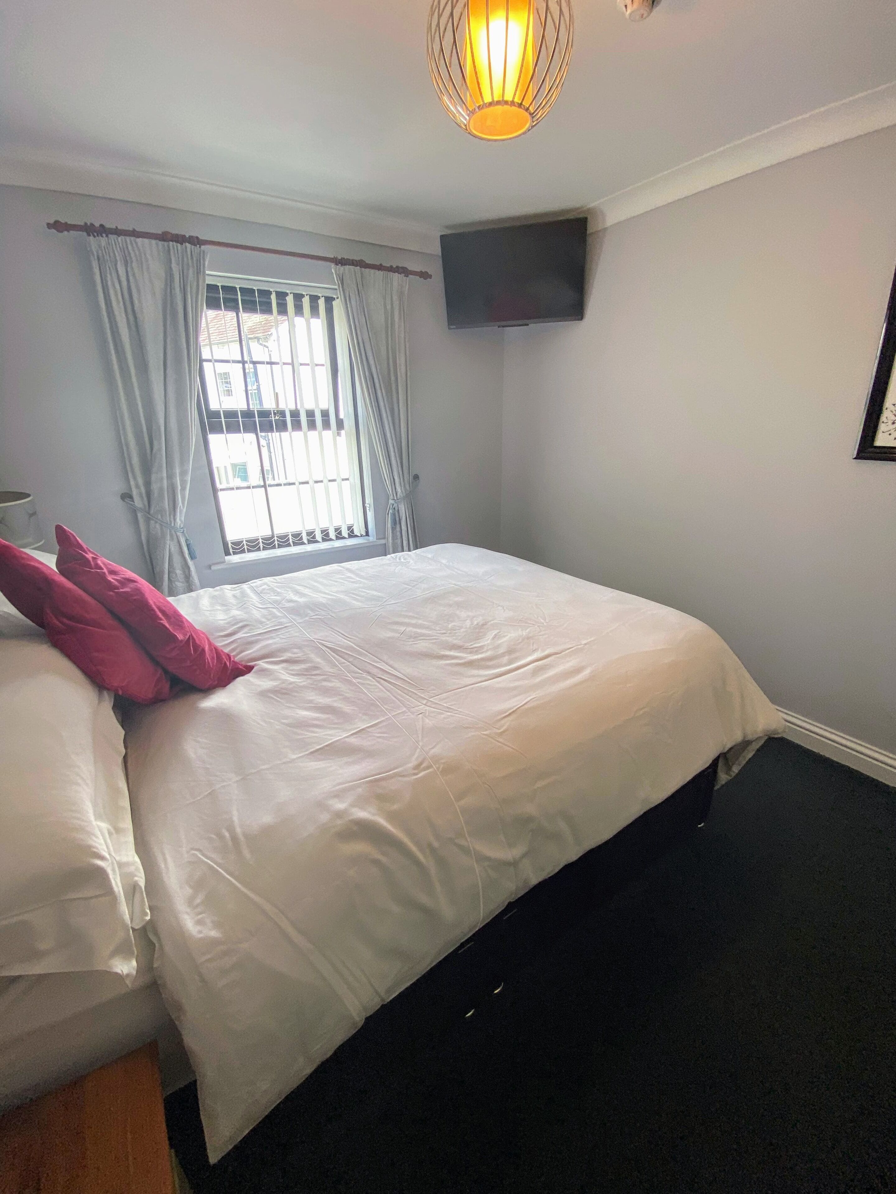 A bedroom with a large bed, a flat-screen TV mounted on the wall, and a window with curtains.