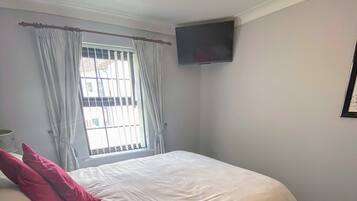 A bedroom with a large bed, a flat-screen TV mounted on the wall, and a window with curtains.