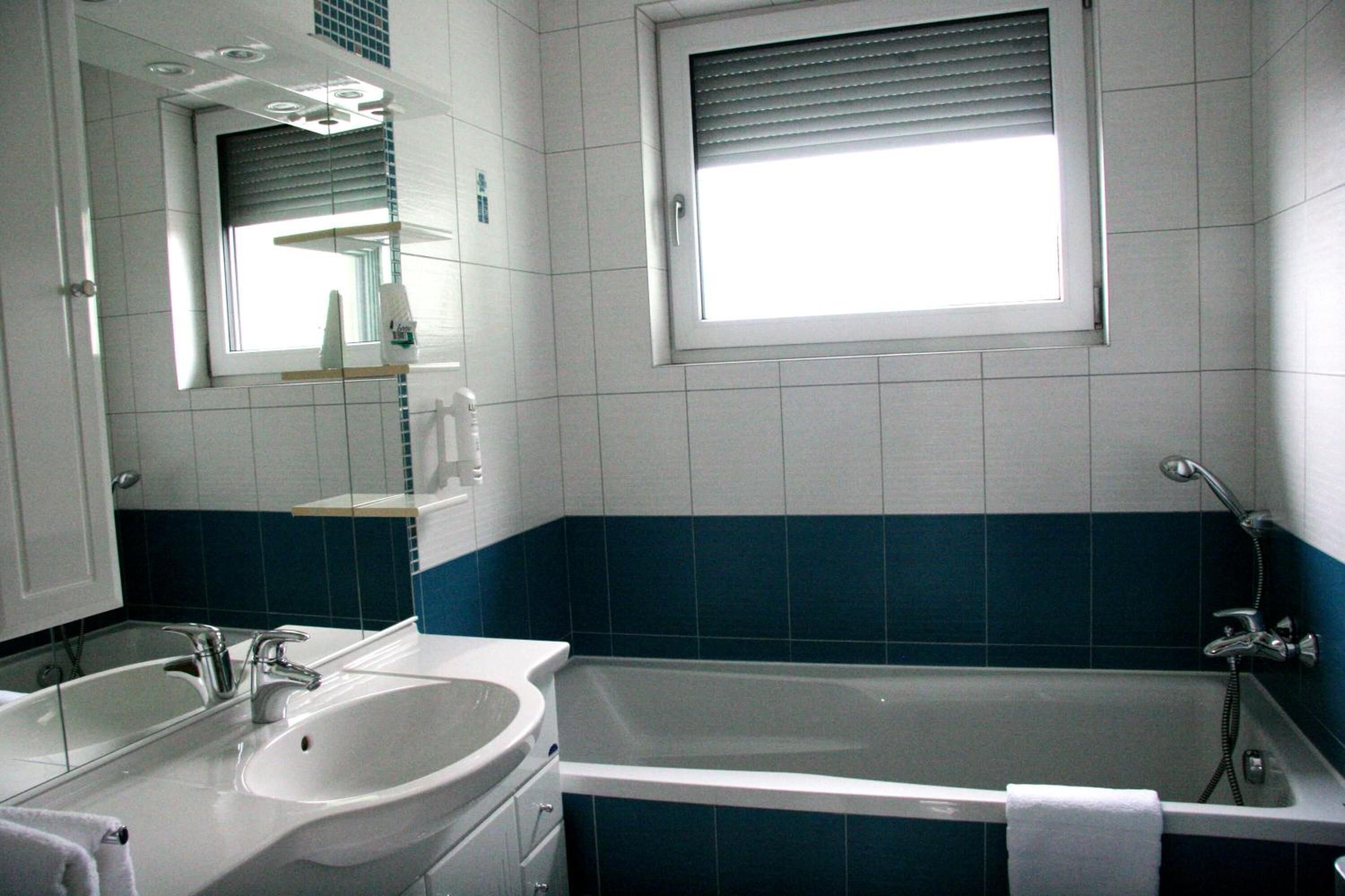 Une salle de bain avec un lavabo, un miroir et une baignoire blancs. Il y a une fenêtre au-dessus de la baignoire et une étagère au-dessus du lavabo.