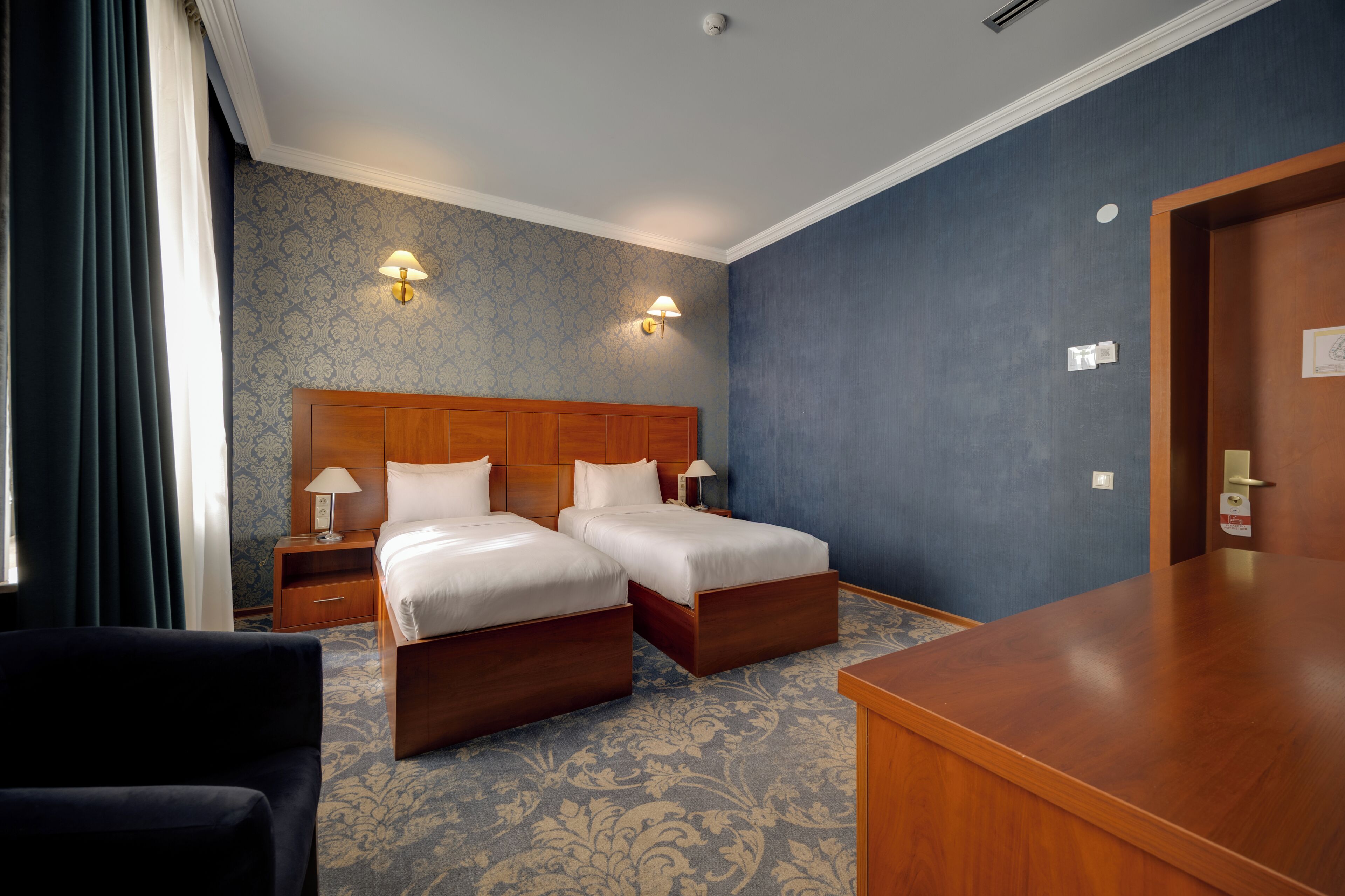 A hotel room with two beds, a wooden headboard, a desk, and a nightstand with lamps.