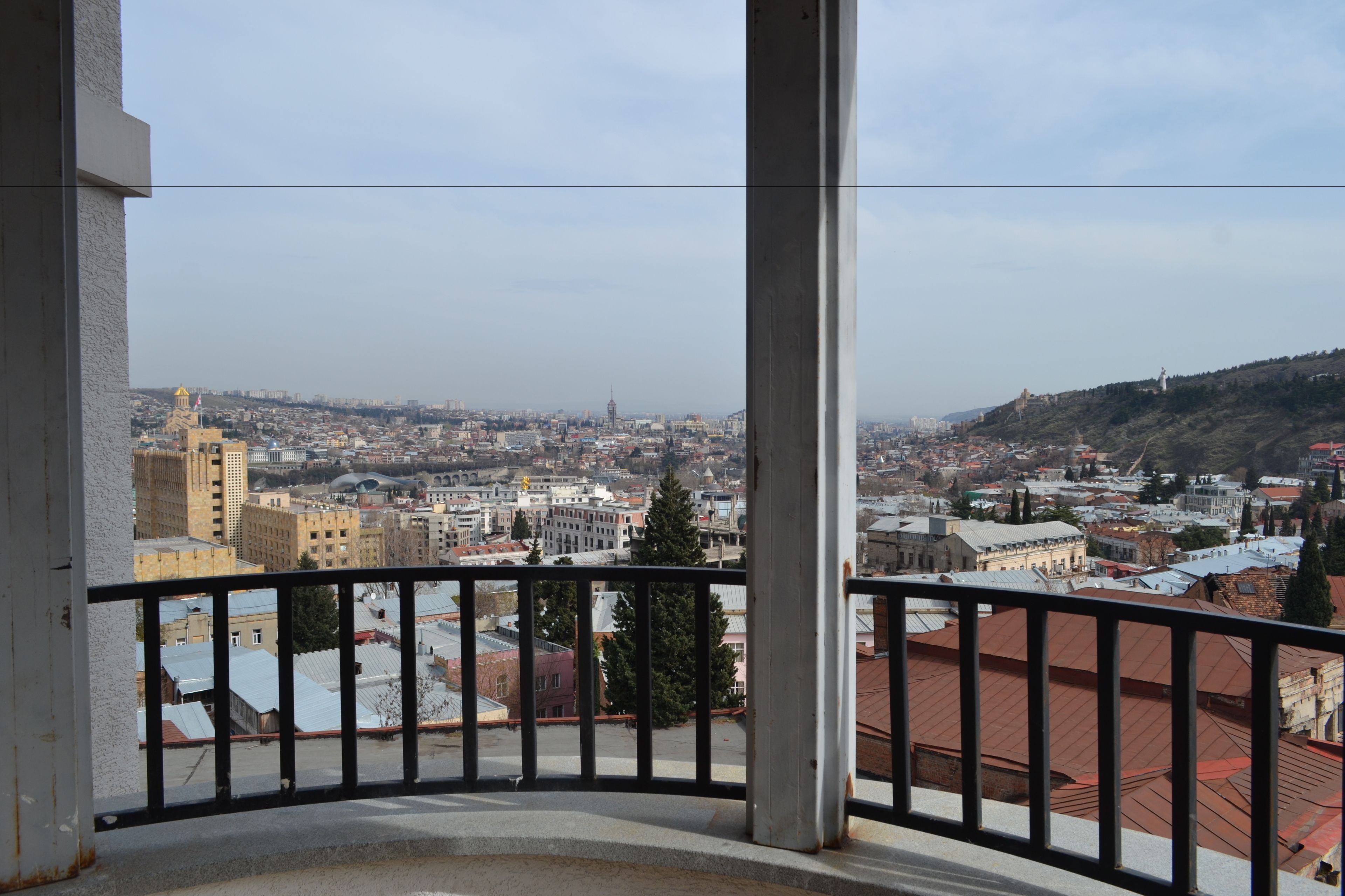 Un balcón con vista a un paisaje urbano, incluyendo edificios y una colina con una estatua.