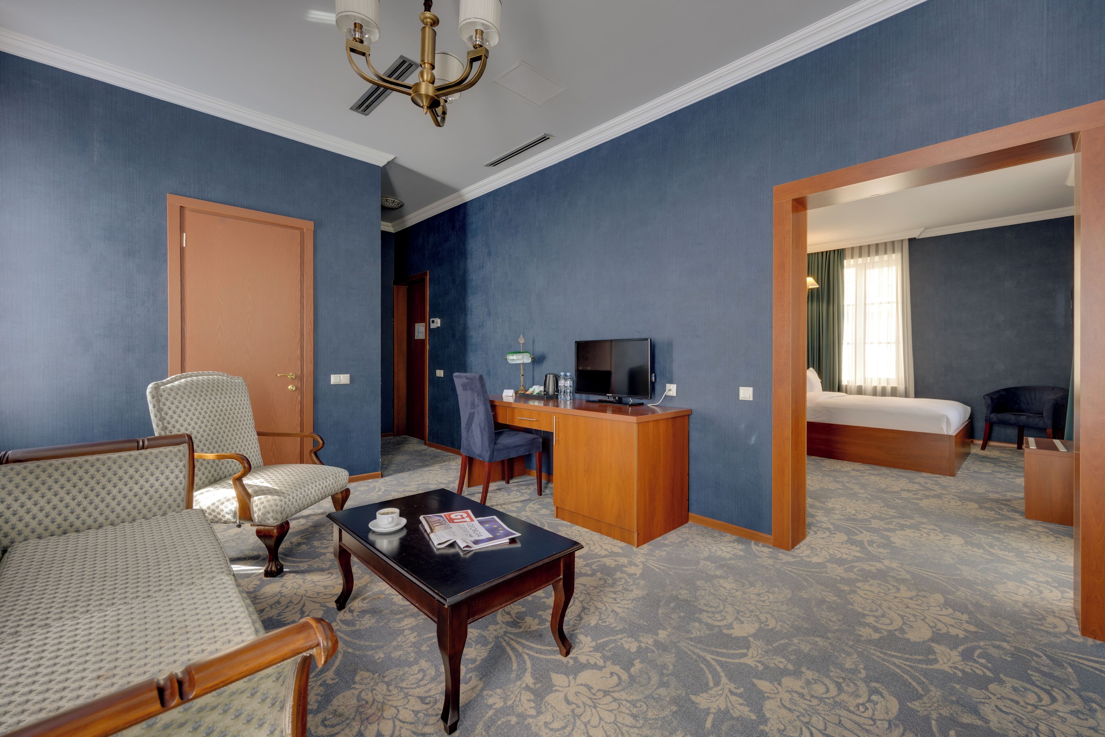 Une chambre d’hôtel comprenant un lit, un bureau avec une télévision, deux fauteuils, une table basse et un sol recouvert de moquette.
