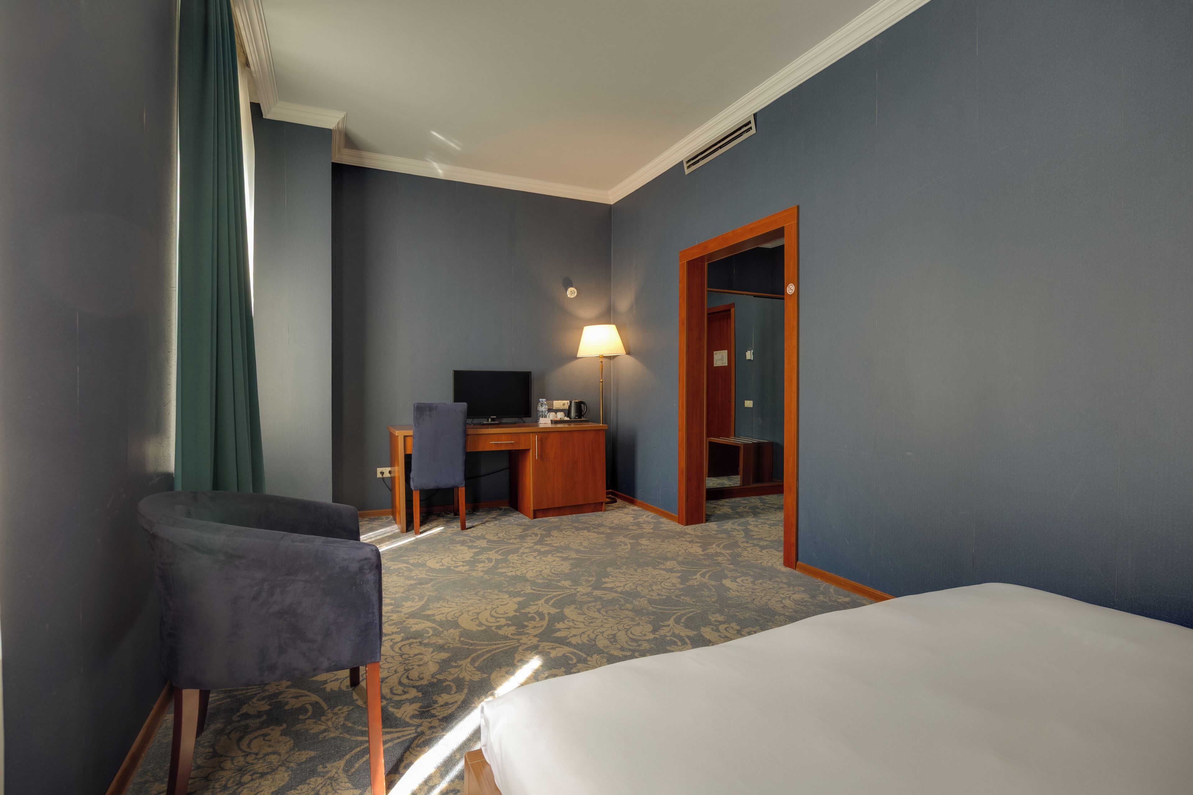 A hotel room with a bed, a desk, a chair, a lamp, and a wardrobe.
