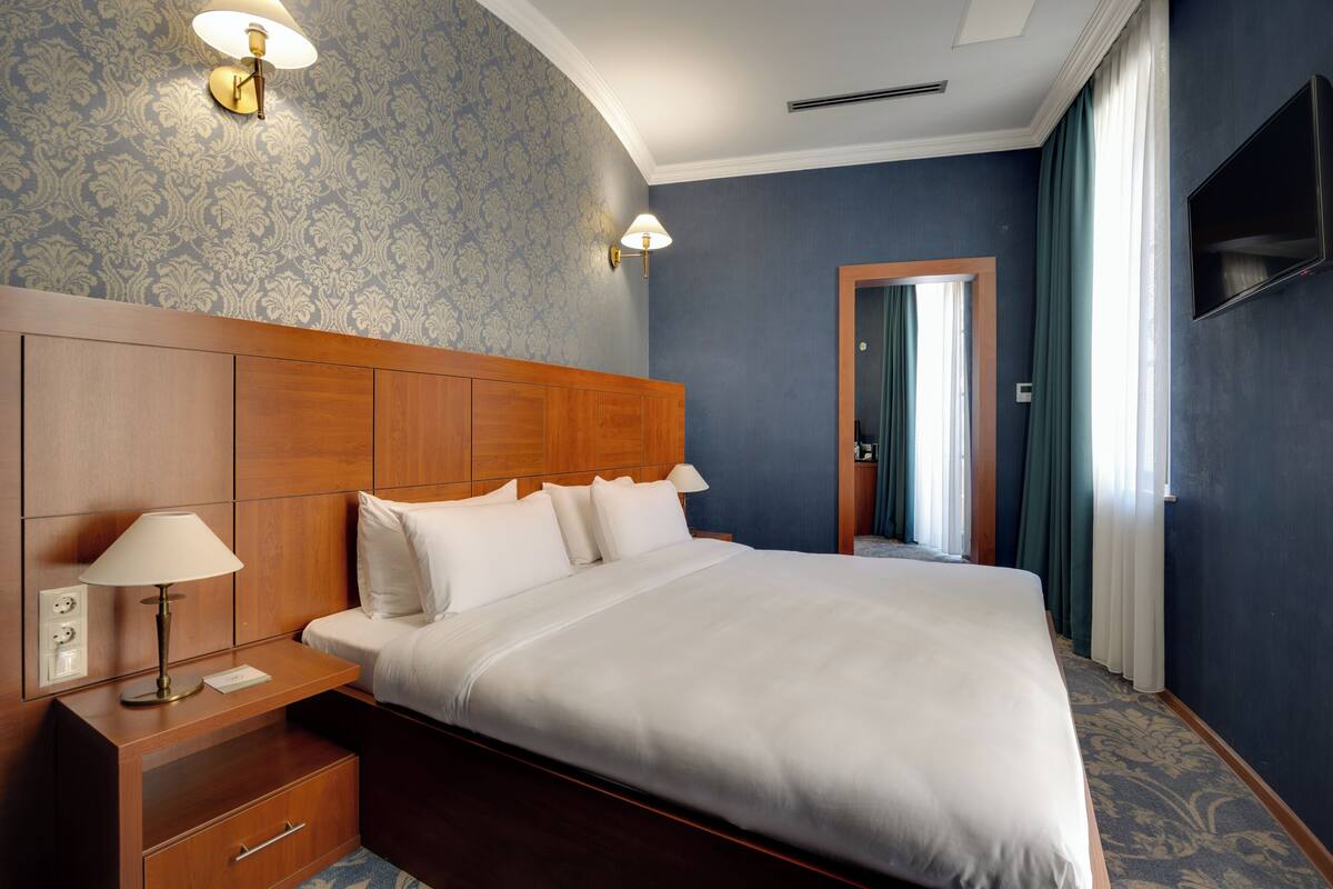 Une chambre d’hôtel avec un grand lit, une t ête de lit en bois, deux lampes de chevet, une table de chevet, un téléviseur fixé au mur et une moquette à motifs.