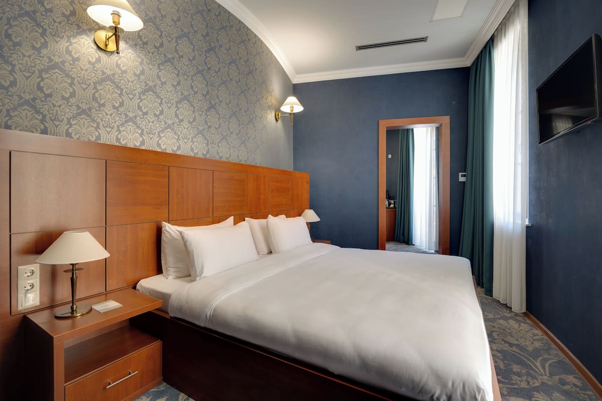A hotel room with a large bed, wooden headboard, two bedside lamps, a nightstand, a TV mounted on the wall, and a patterned carpet.