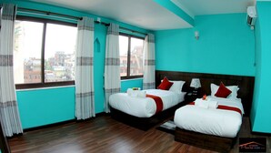 A hotel room with two beds, teal walls, wooden flooring, and a view of buildings through large windows.