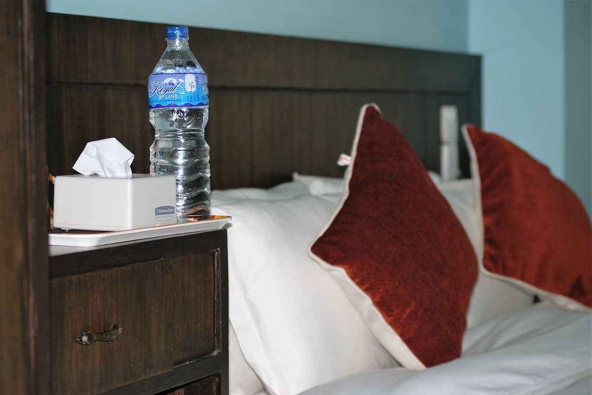 A hotel room with a wooden headboard, a bedside table with a tissue box and a bottle of water, and a red throw pillow on the bed.