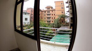A view through a window with a black frame, revealing a balcony with a metal railing and a green area, possibly a garden or courtyard, with buildings in the background.