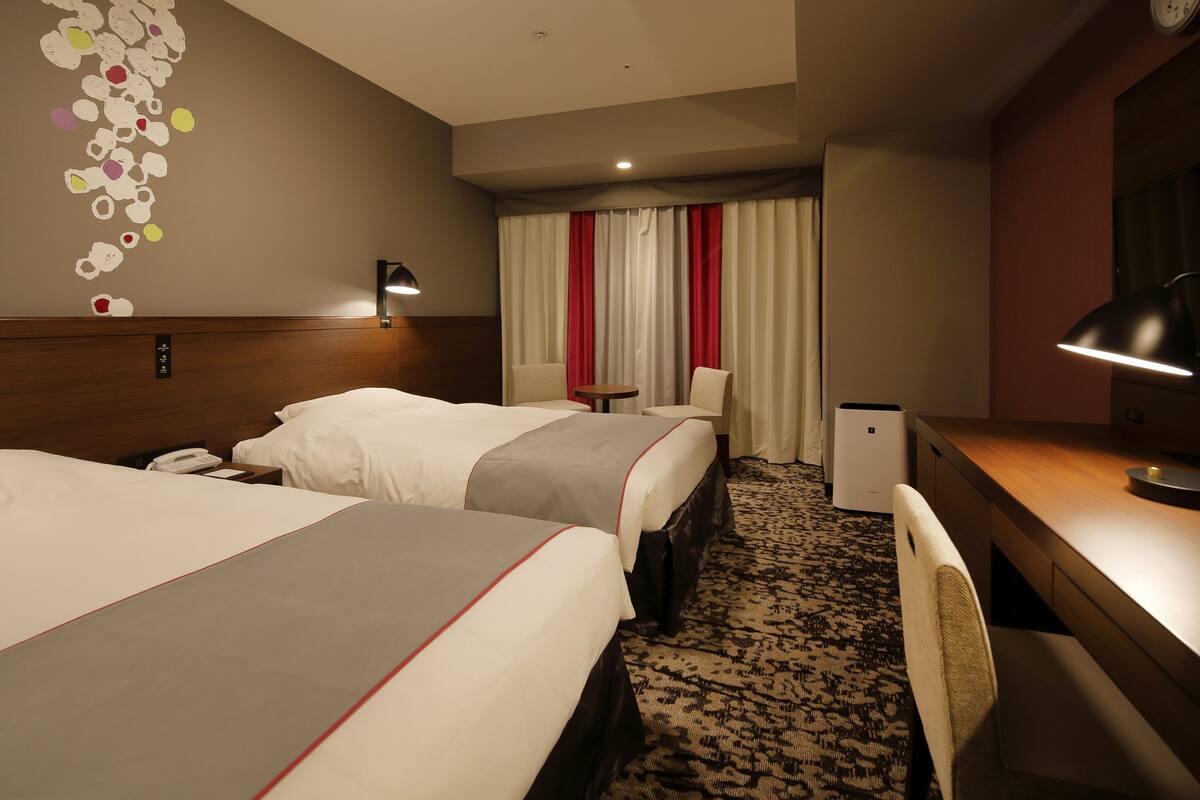 Une chambre d’hôtel avec deux lits, un bureau, une chaise, une lampe, une horloge et un mur orné d’un motif décoratif.