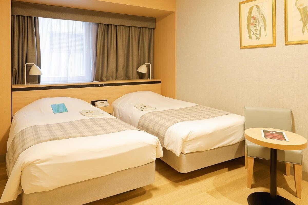 Une chambre d’hôtel avec deux lits, une petite table et une chaise. Il y a deux lampes sur les tables de chevet, une fenêtre avec des rideaux et deux reproductions encadrées de motifs botaniques au mur.