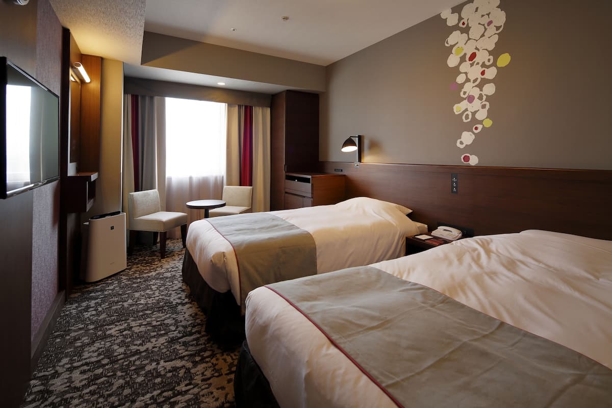 Une chambre d’hôtel avec deux lits, un bureau, une chaise, une petite table, une lampe et un élément décoratif mural.