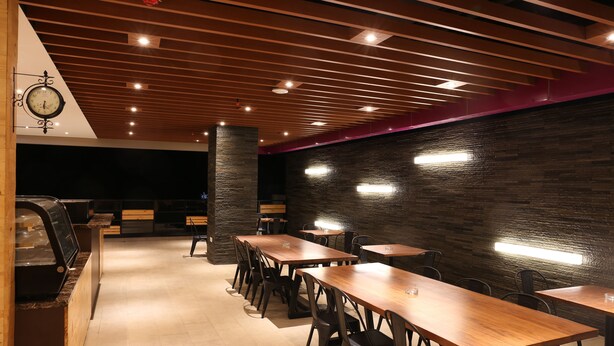 A modern restaurant interior with wooden tables and chairs, a clock on the wall, and a stone pillar. - Grand Citihub Hotel @kajoetangan (Malang)