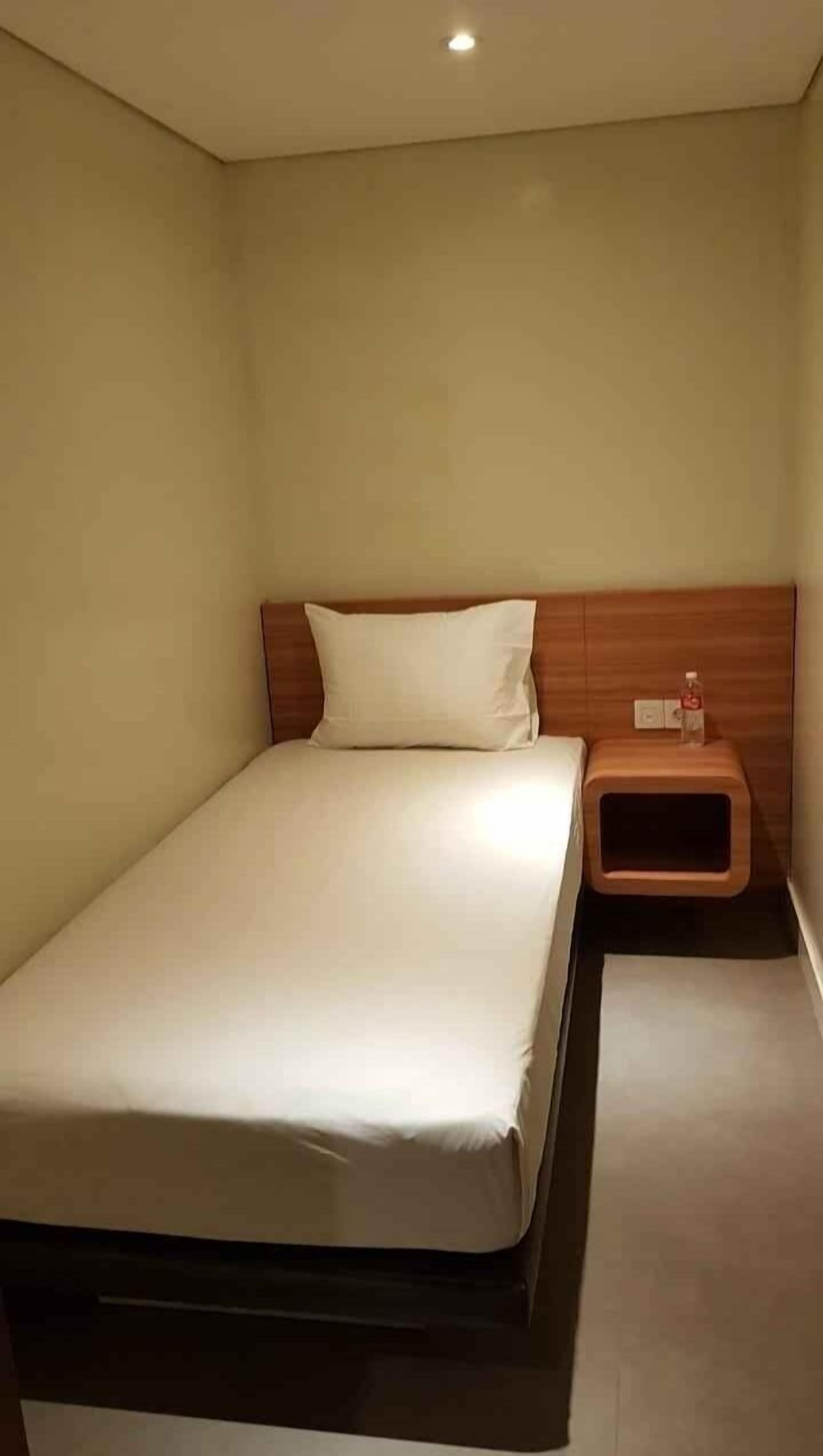 Un lit simple avec une tête de lit en bois, une table de chevet avec une bouteille et un interrupteur sur le mur.