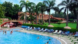 A swimming pool with lounge chairs and umbrellas, surrounded by palm trees and a building with arches. - Ashok Country Resort (New Delhi)