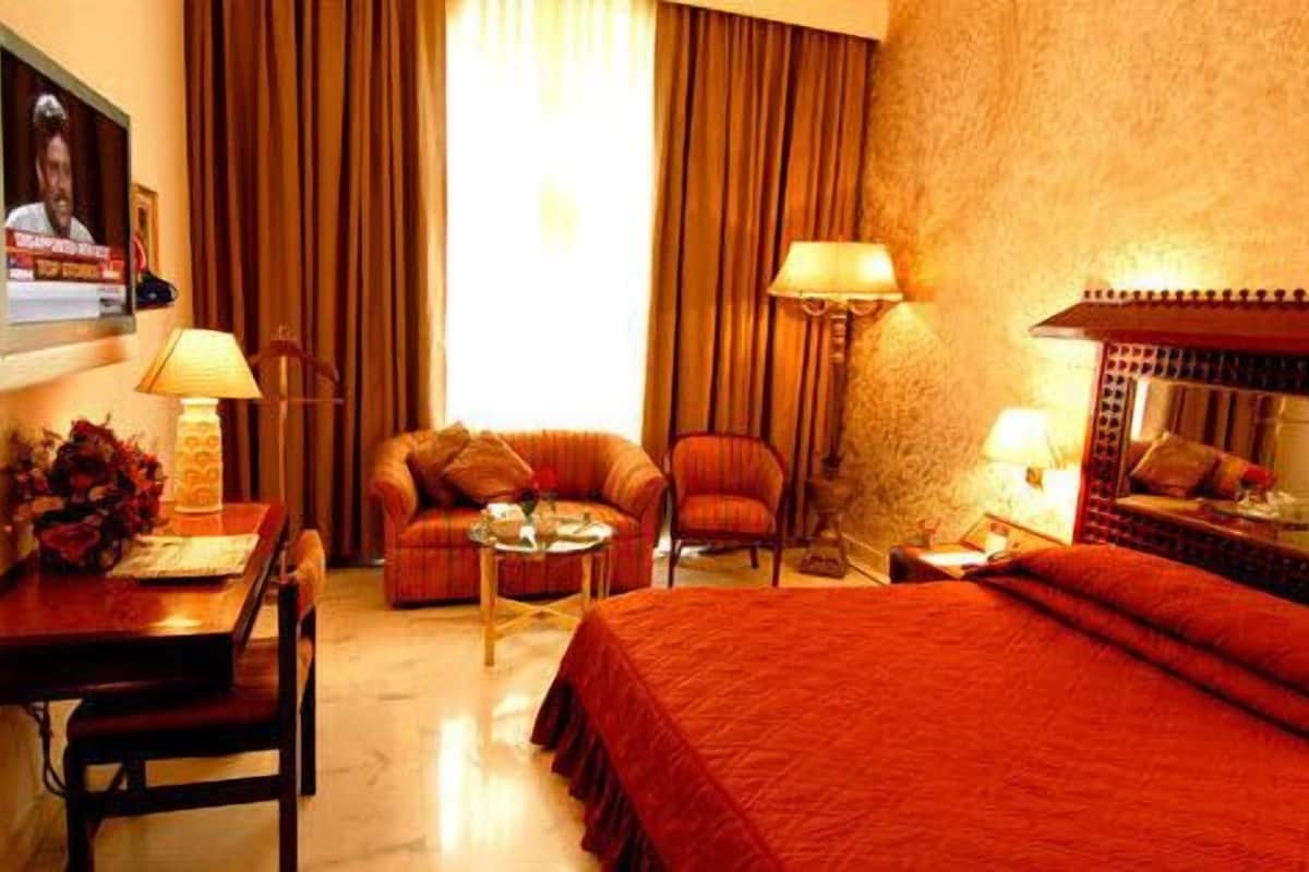 A hotel room with a bed, two armchairs, a small table, and a desk. There is a television on the wall and a lamp on the desk.