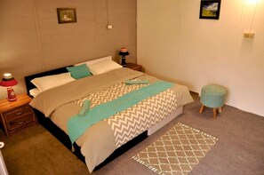 Una cama bien hecha con una colcha estampada, dos lámparas de noche, un taburete de madera y una alfombra en el suelo.