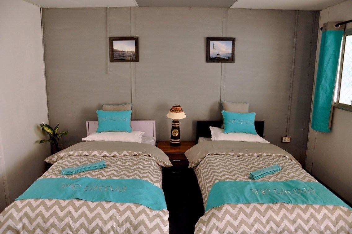 A hotel room with two beds, each with a teal and white bedspread, a central lamp, and framed pictures on the wall.