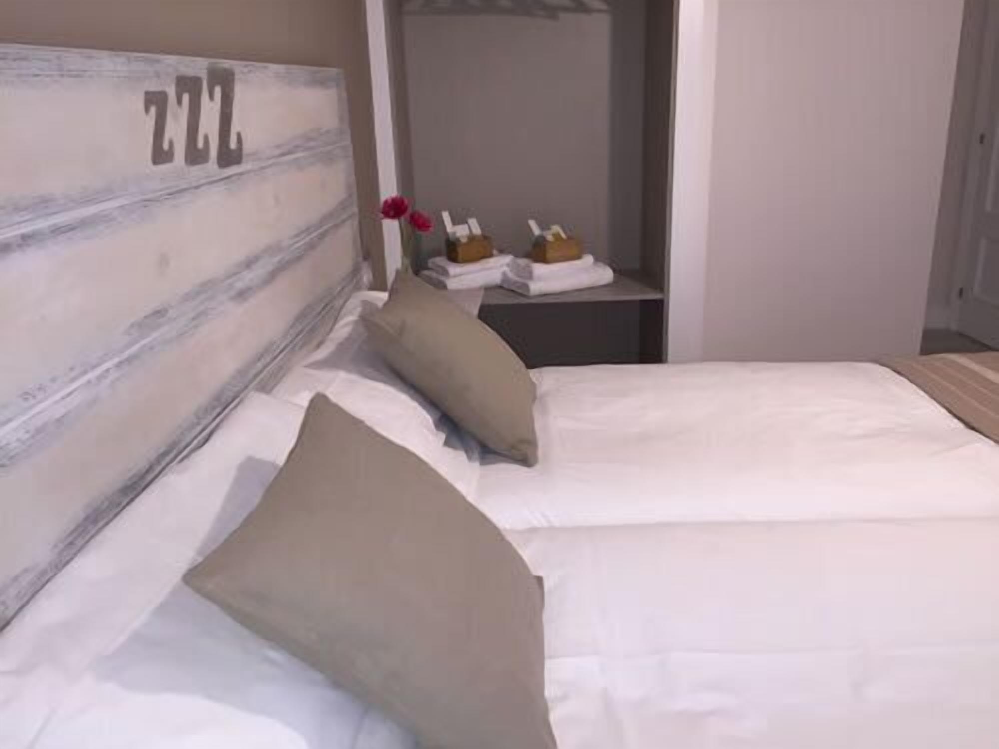 Una cama bien hecha con ropa de cama blanca y beige, un cabecero de madera con el número '222', y una mesita de noche con un florero de flores rojas y un par de zapatos.