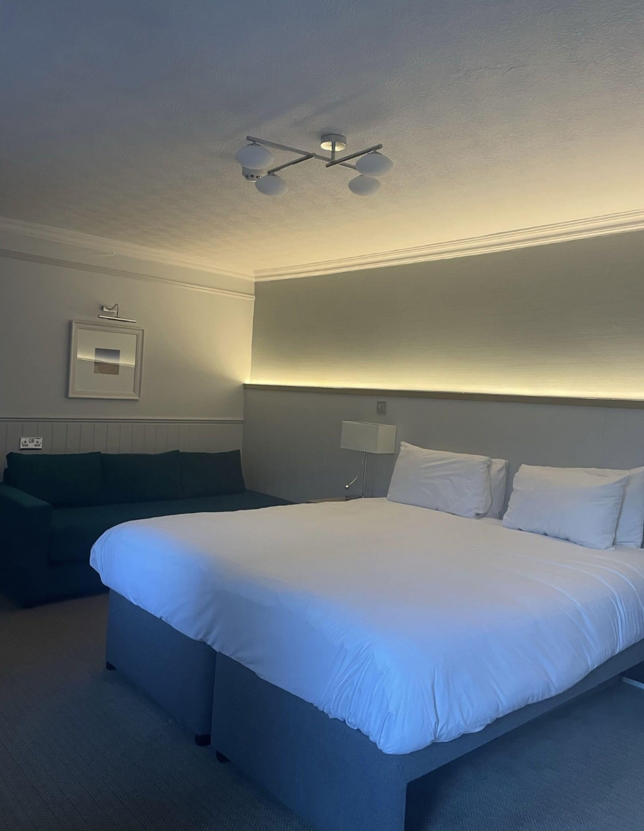 A hotel room with a large bed, a green sofa, a ceiling light fixture, and a wall-mounted lamp.