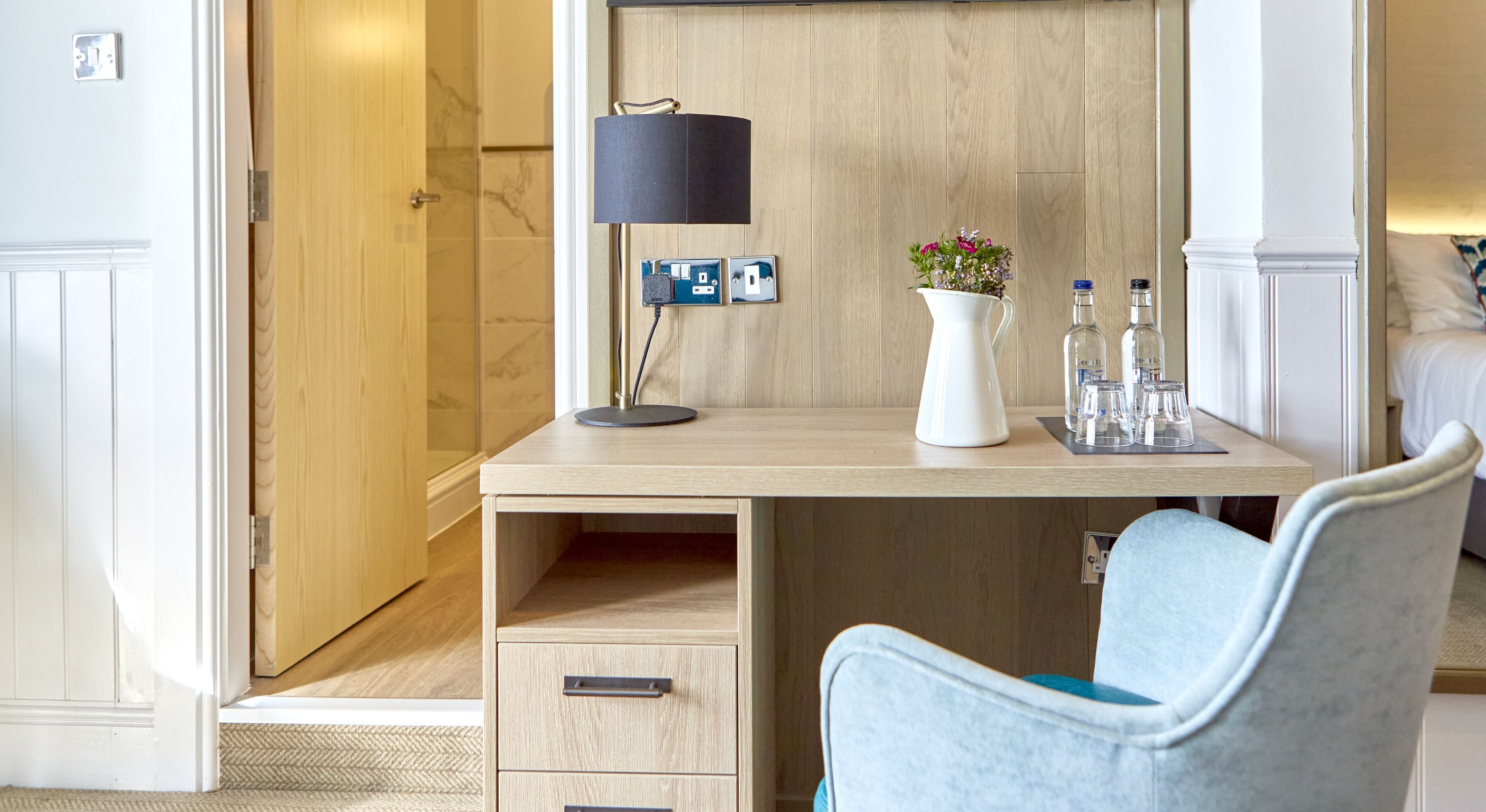 Ein Hotelzimmer mit einem Holztisch, einer Lampe, einer Vase mit Blumen und einem Sessel.