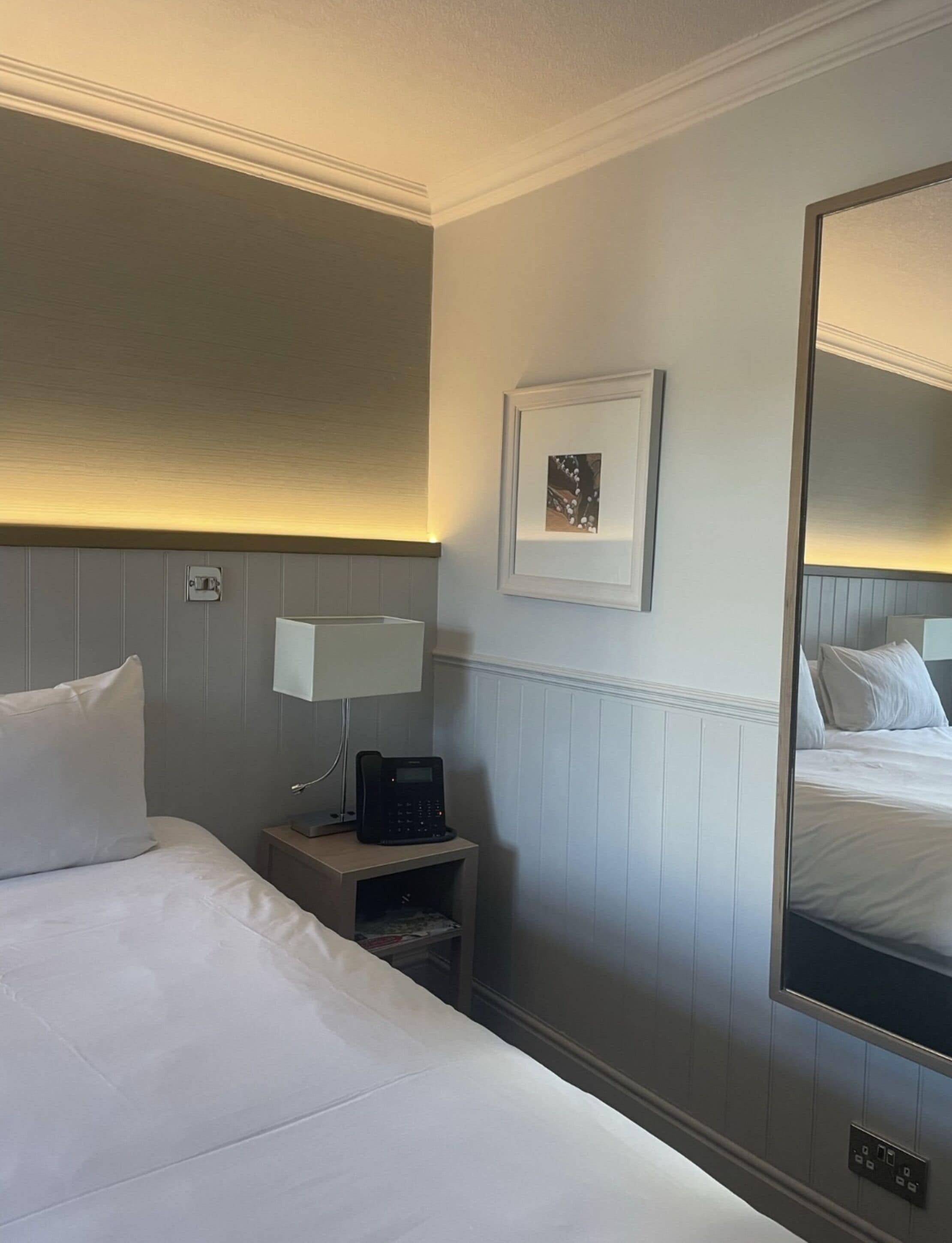 A hotel room with a neatly made bed, a bedside table with a lamp, a phone, and a framed picture on the wall.