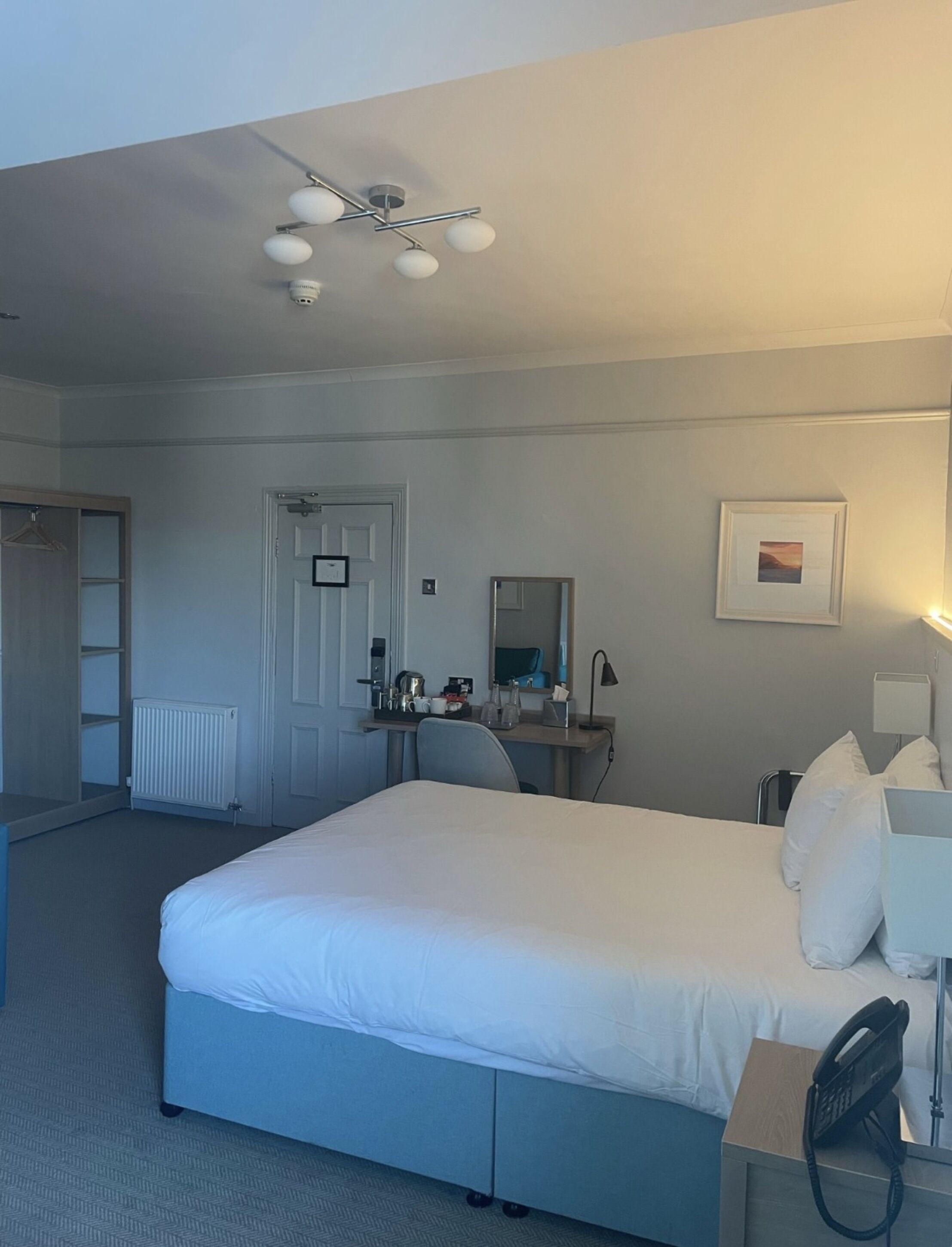 A hotel room with a large bed, a desk with a chair, a radiator, a door, a mirror, and a framed picture on the wall.