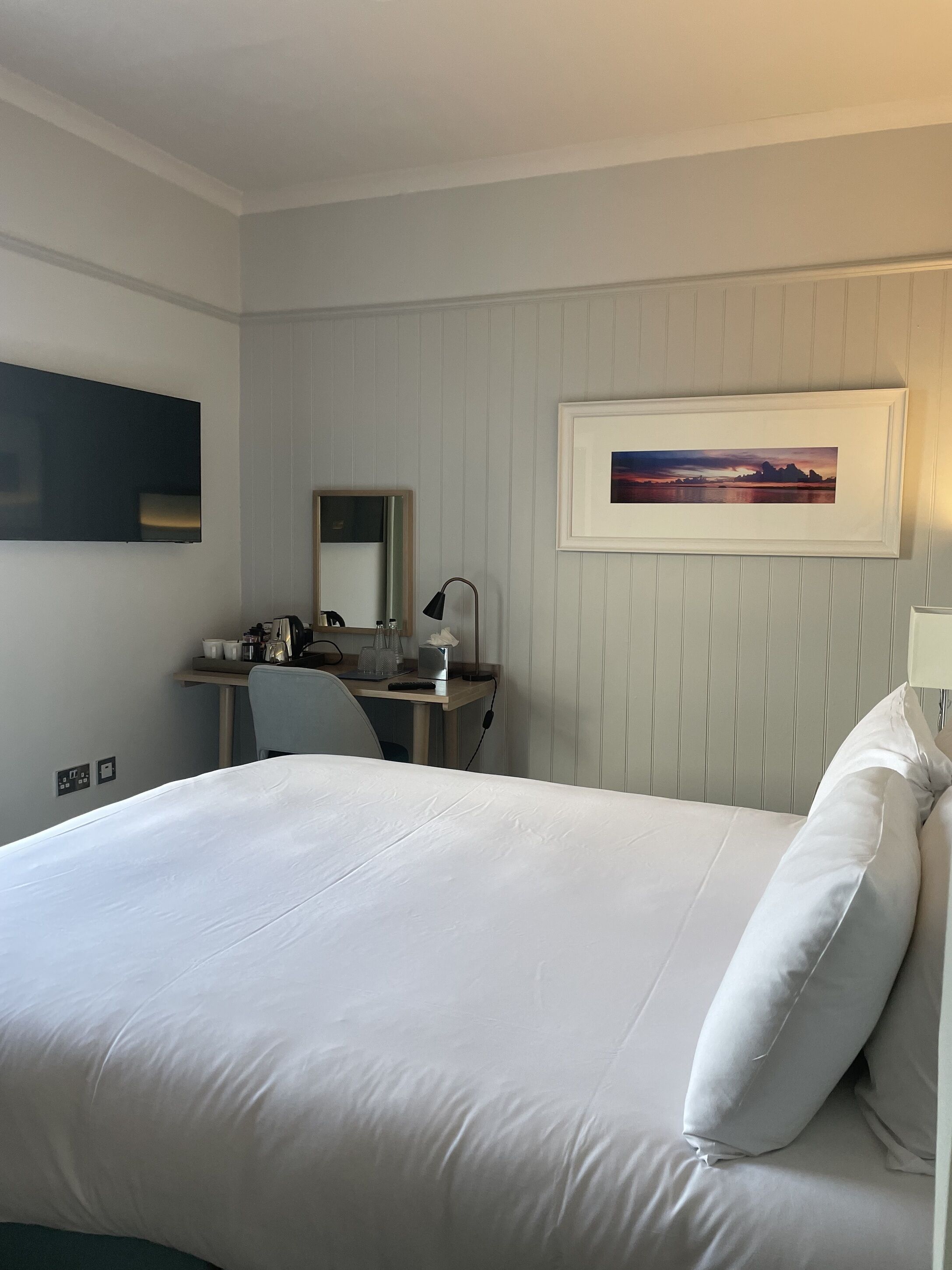 Habitación de hotel con una cama grande, un escritorio con una silla, un televisor de pantalla plana, un cuadro en la pared y una mesita de noche con lámpara.