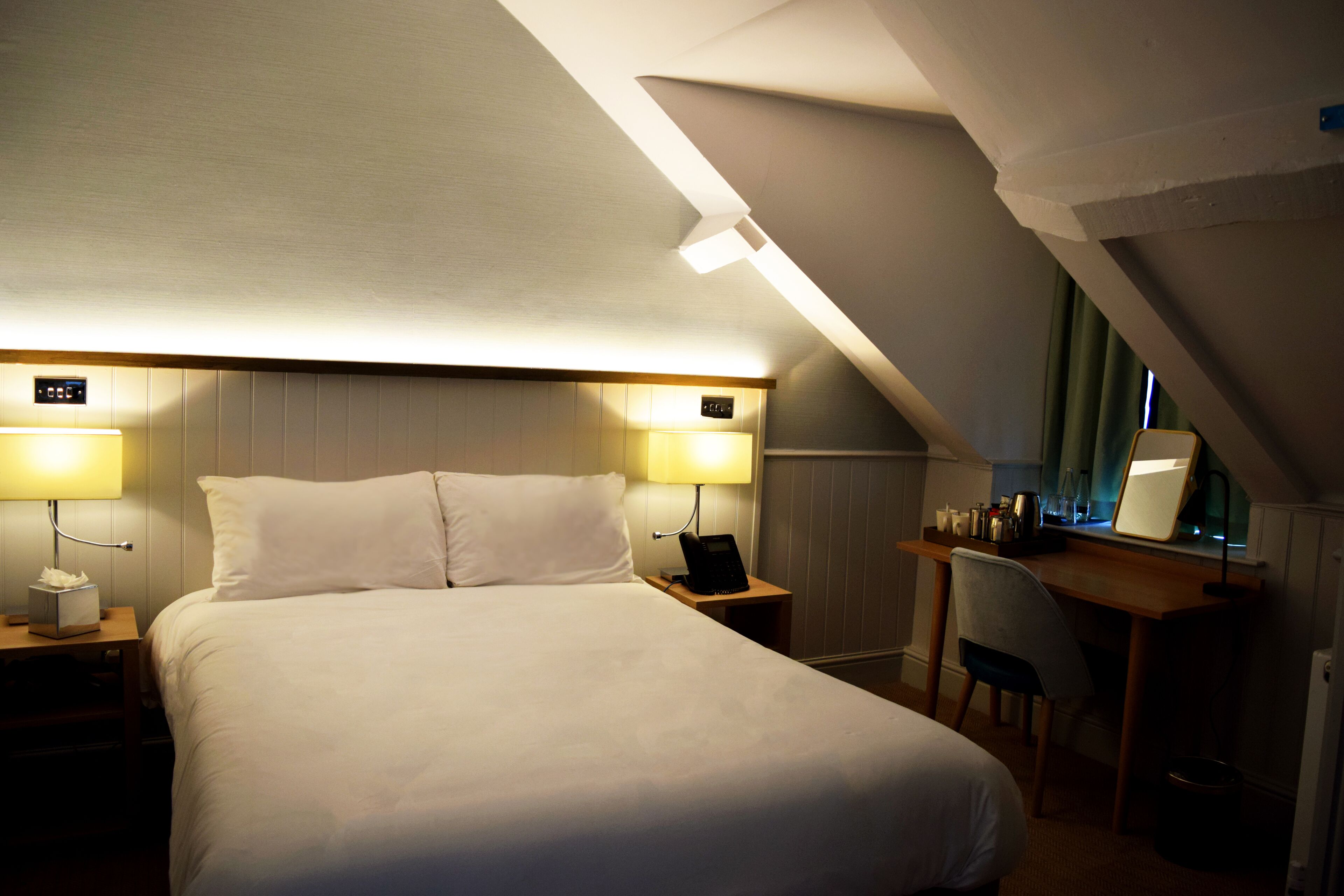 Una habitación de hotel con una cama grande, dos lámparas de noche, un escritorio con una silla y una mesita con un teléfono y una lámpara.