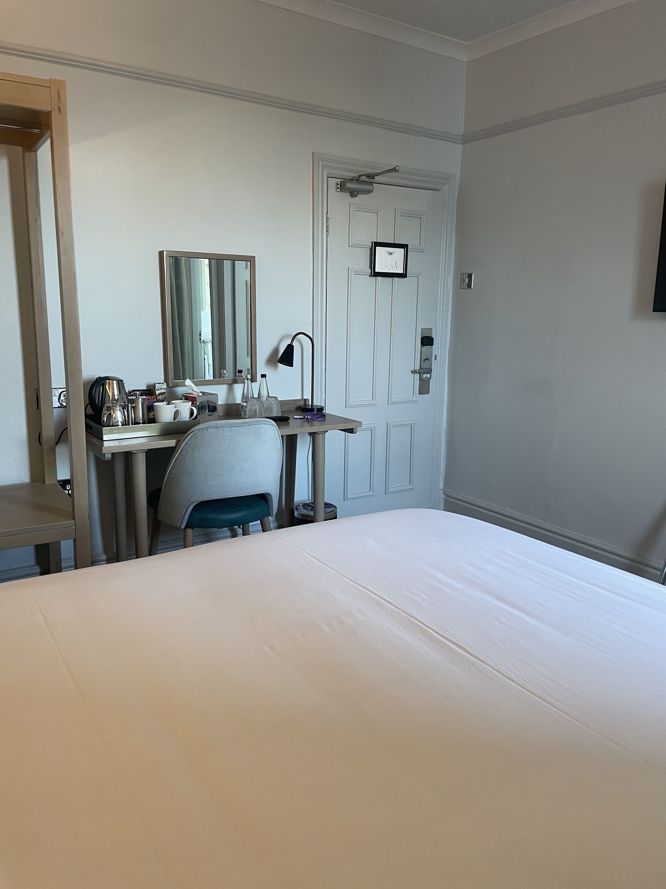 Habitació d'hotel amb llit, escriptori, cadira i porta amb interruptor de llum i un quadre.