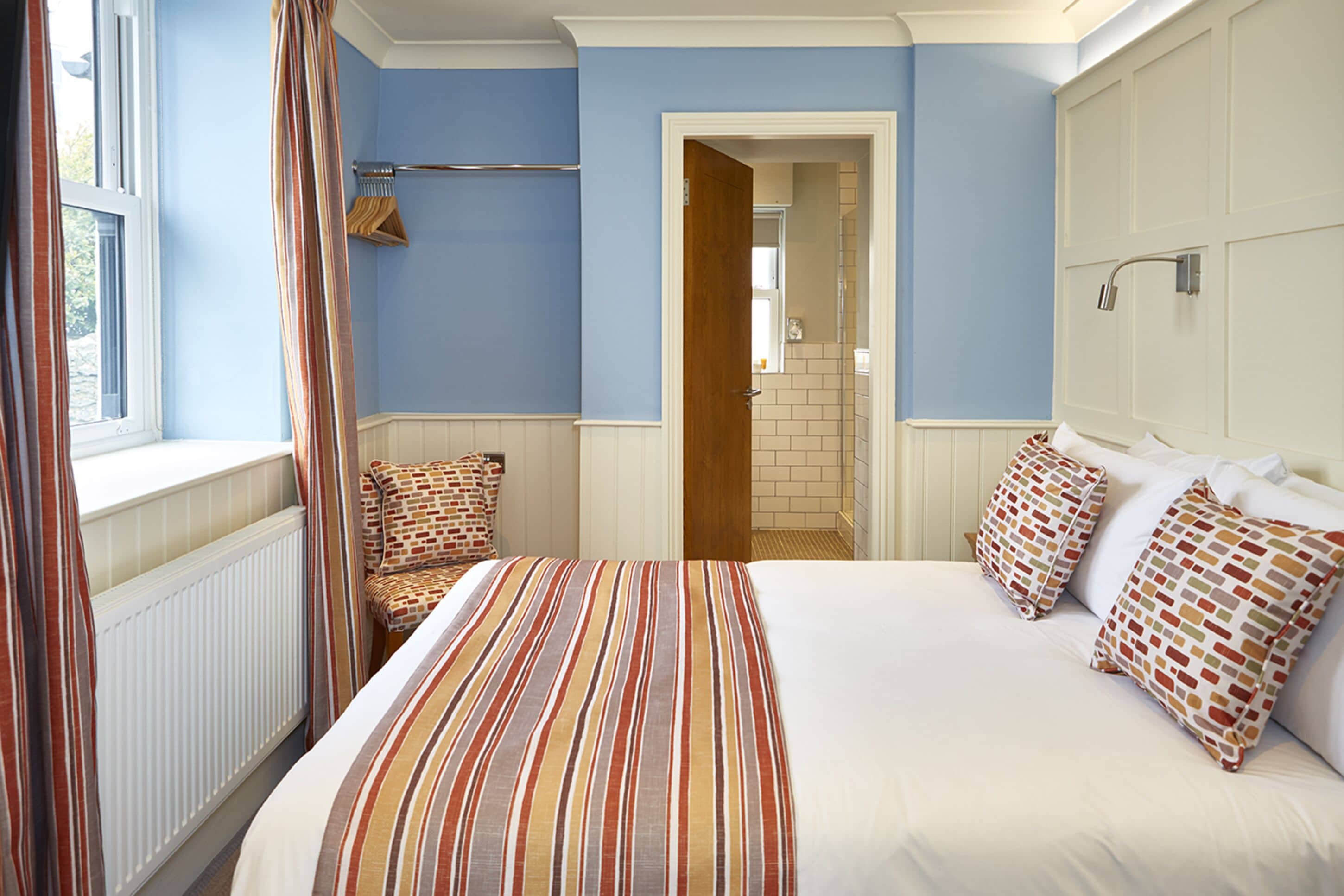 Une chambre à coucher avec un grand lit, deux oreillers, une fenêtre avec des rideaux, un radiateur et une porte donnant sur une salle de bain.