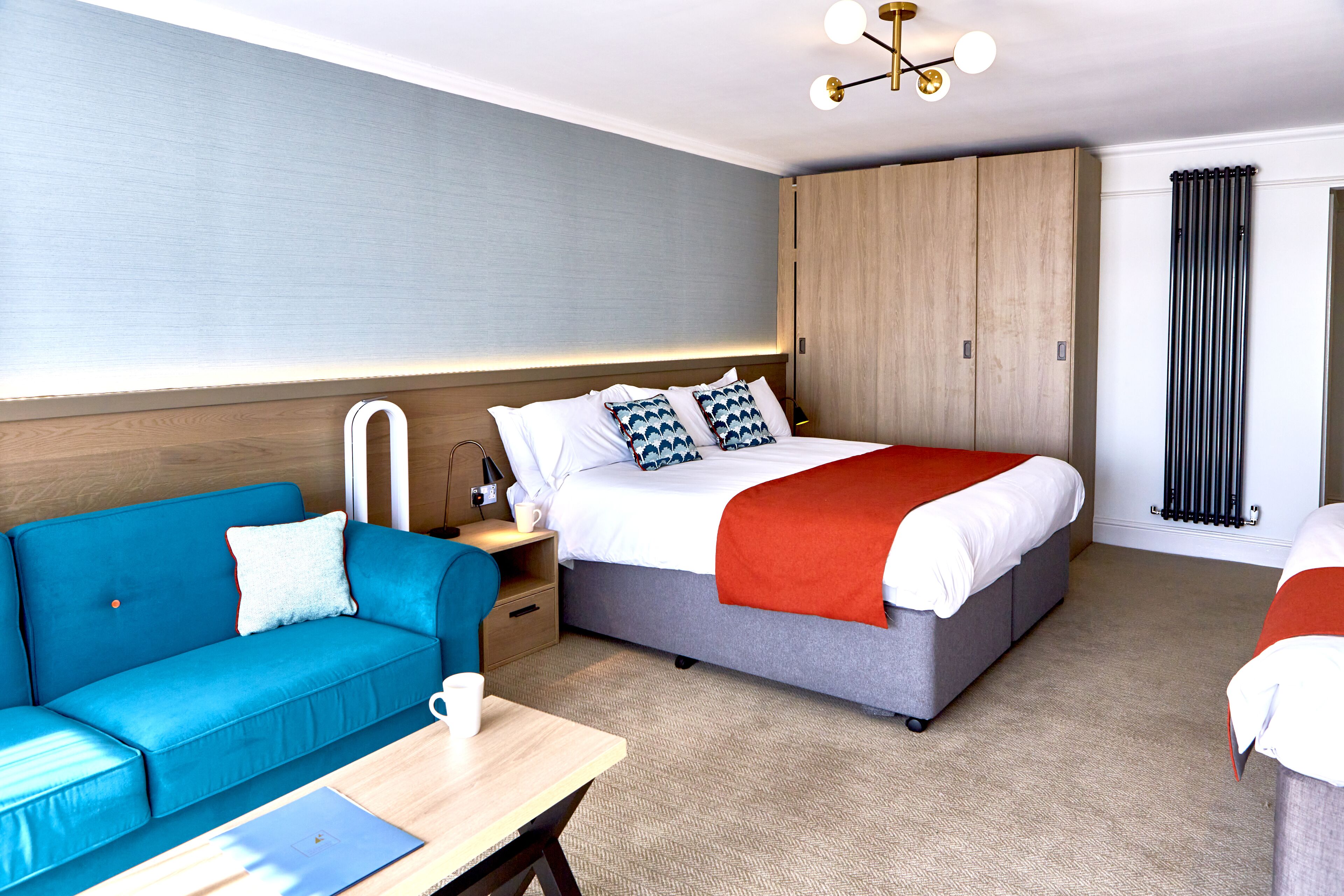 A modern hotel room with a bed, a blue sofa, a wooden wardrobe, and a desk with a laptop.