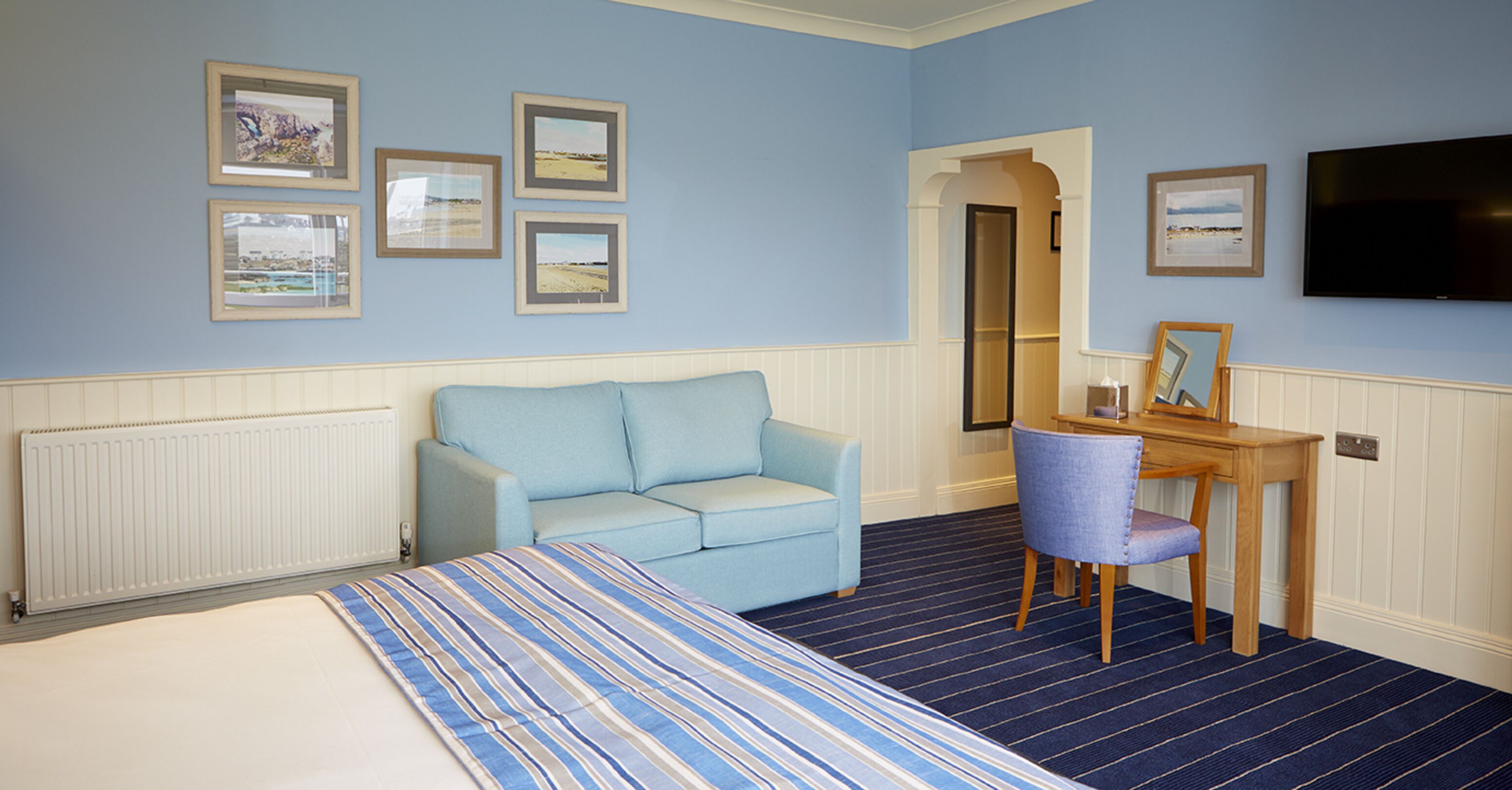 A room with a bed, a light blue sofa, a wooden desk, and a TV on the wall.