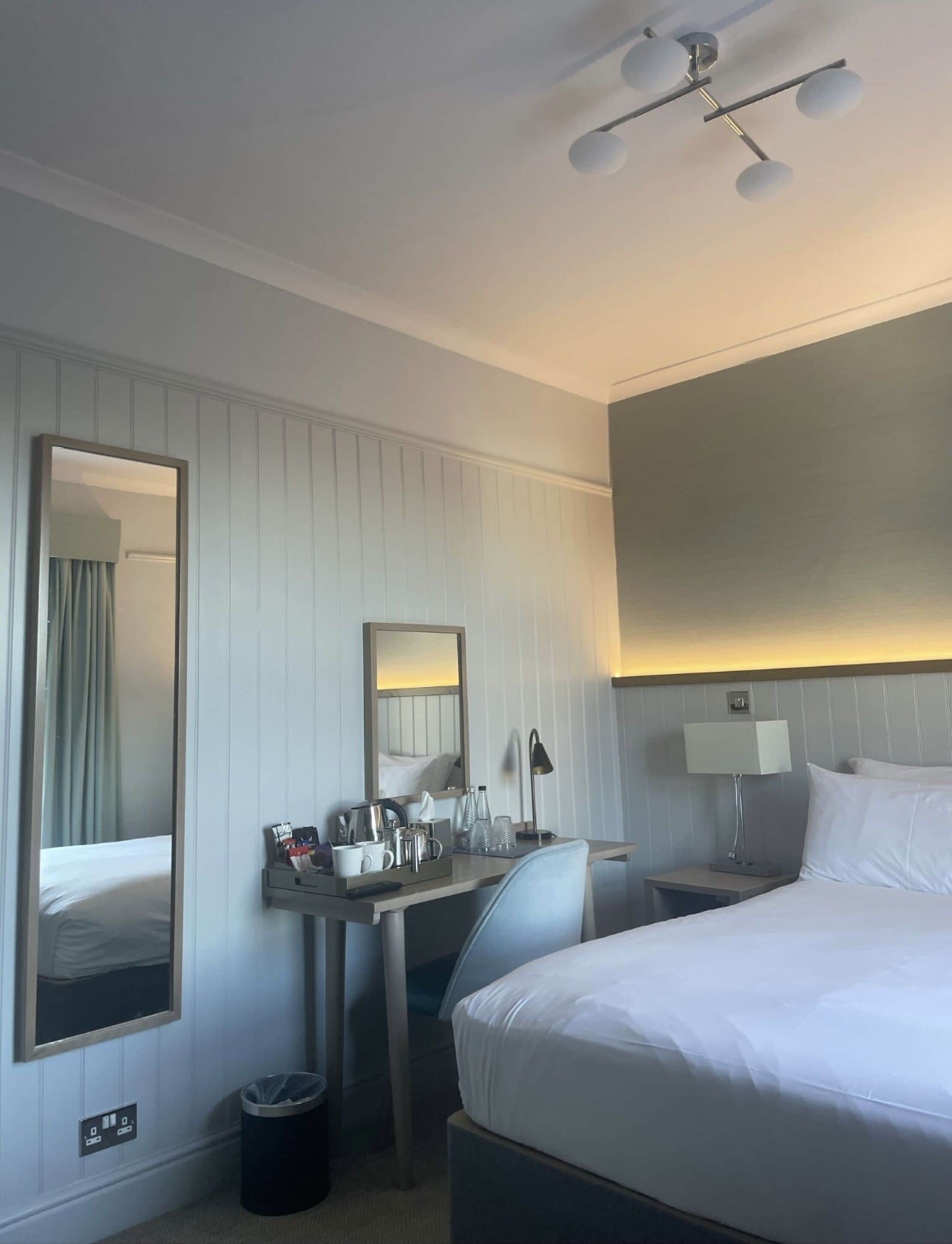 A hotel room with a bed, a desk with a mirror, a lamp, and a trash bin.