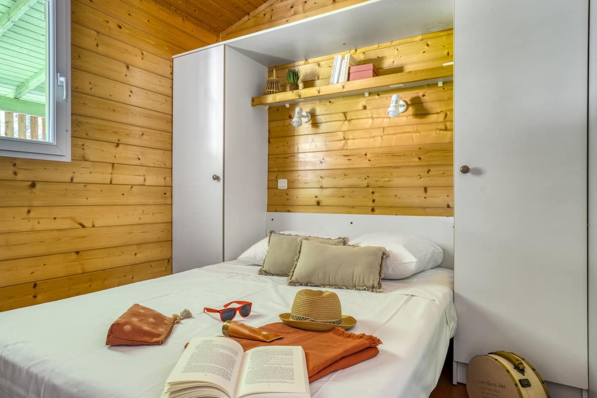Uma cama bem arrumada com almofadas, um chapéu, óculos de sol, um livro e uma mala em um quarto com paredes de madeira e um guarda-roupa branco.
