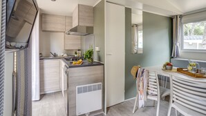 A modern kitchen with a built-in stove, a microwave, and a ventilation hood. There is a dining area with a table and chairs, and a window with curtains. - Camping Le Bois Fleuri (Argelès-sur-Mer)