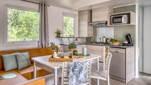 A modern kitchen with a built-in microwave, a coffee maker, and a dining area with a white table and chairs. - Camping Le Bois Fleuri (Argelès-sur-Mer)