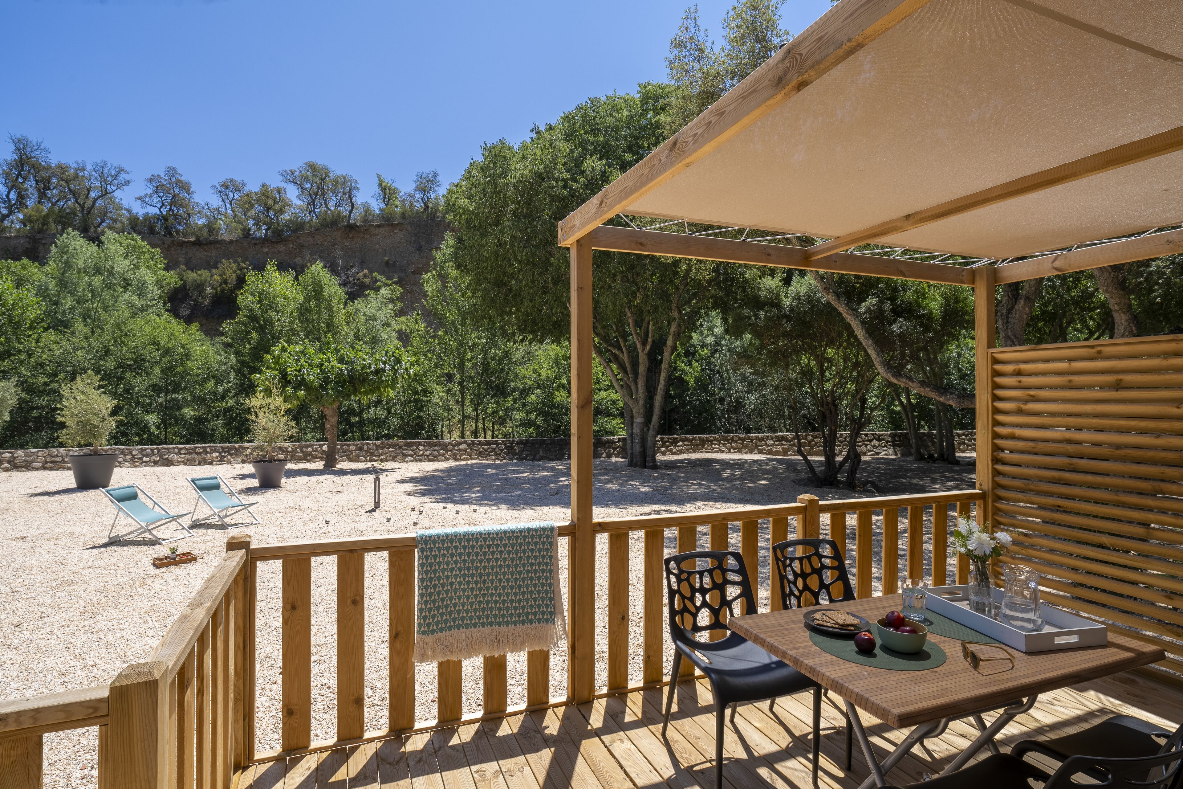 A wooden deck with a table and chairs, a covered seating area, and a view of a sandy area with lounge chairs.