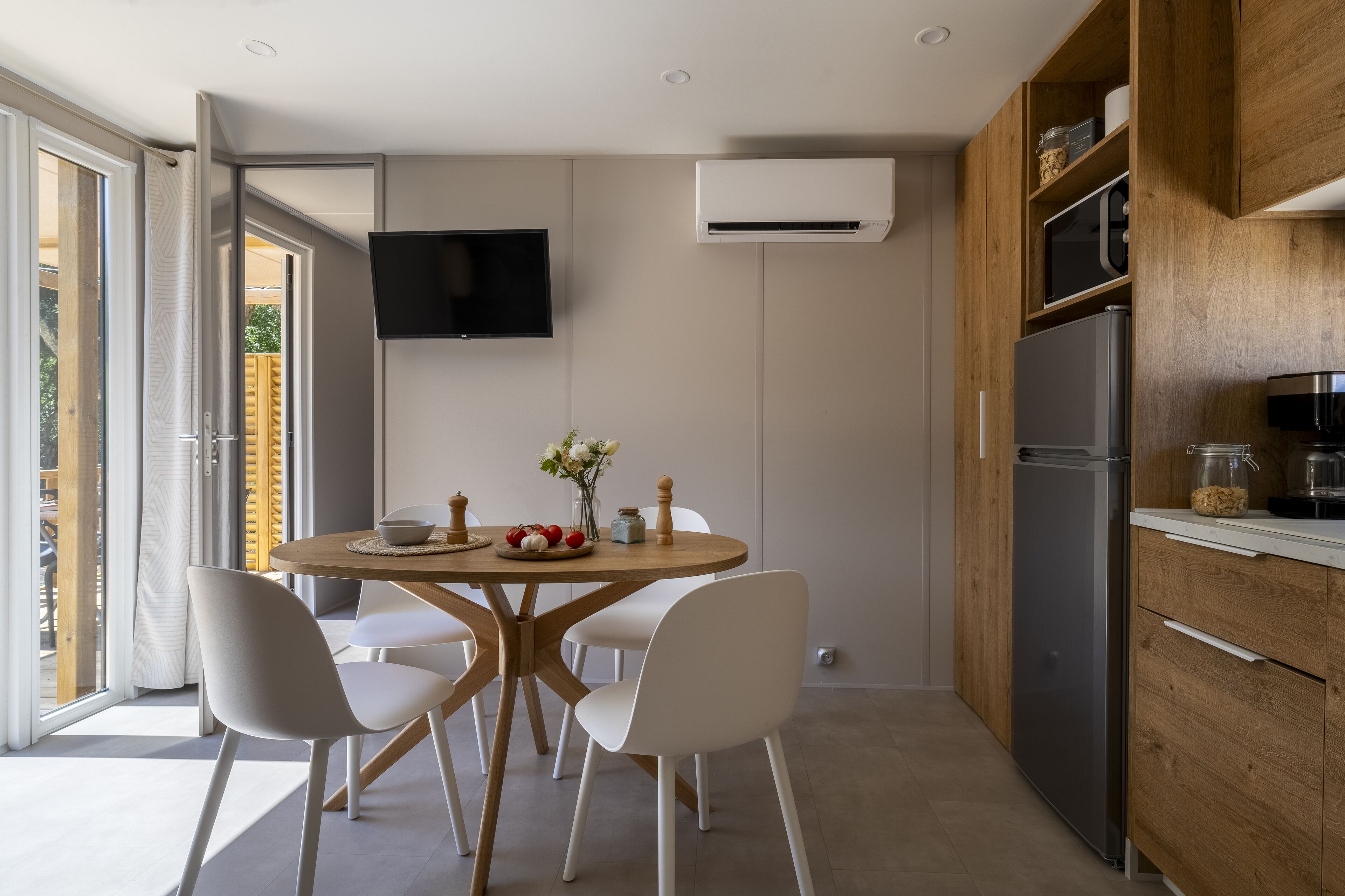 A modern kitchen with a dining area, featuring a round wooden table, white chairs, a flat-screen TV, and built-in appliances.