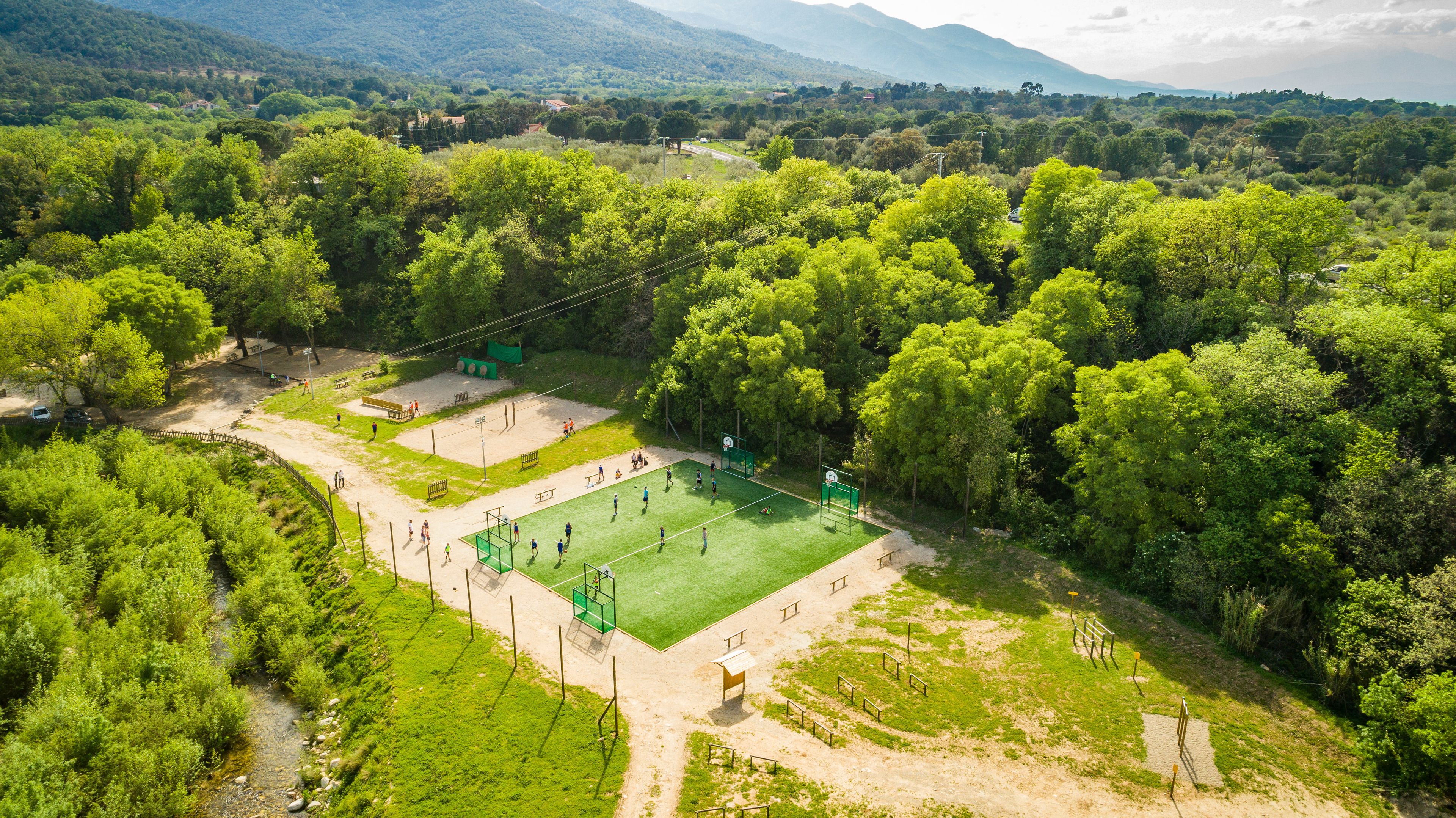 An aerial view of a park with a basketball court surrounded by dense trees and a mountainous backdrop.