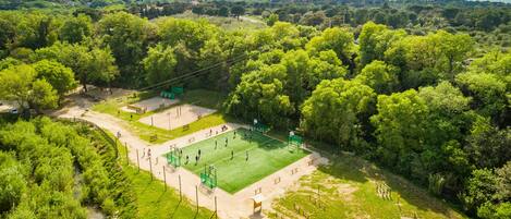An aerial view of a park with a basketball court surrounded by dense trees and a mountainous backdrop.