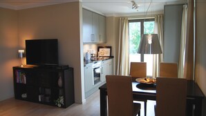 Una sala de estar moderna con un televisor de pantalla plana, una estantería, una mesa de comedor con sillas, una zona de cocina con armarios y una ventana con cortinas.