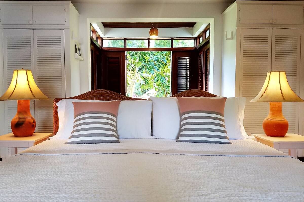 A bedroom with a large bed, two bedside lamps, and a window with a view of greenery.