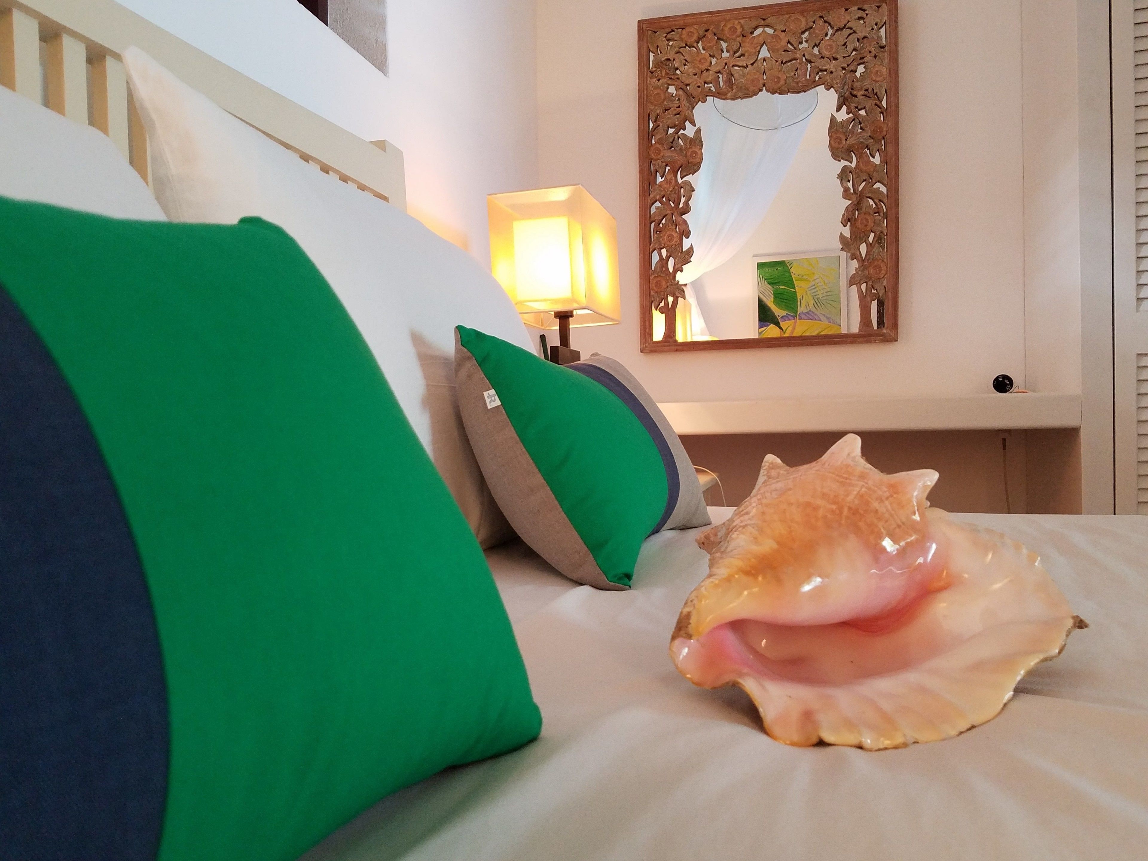A bed with green and beige pillows, a large seashell, a wooden mirror, and a lamp.