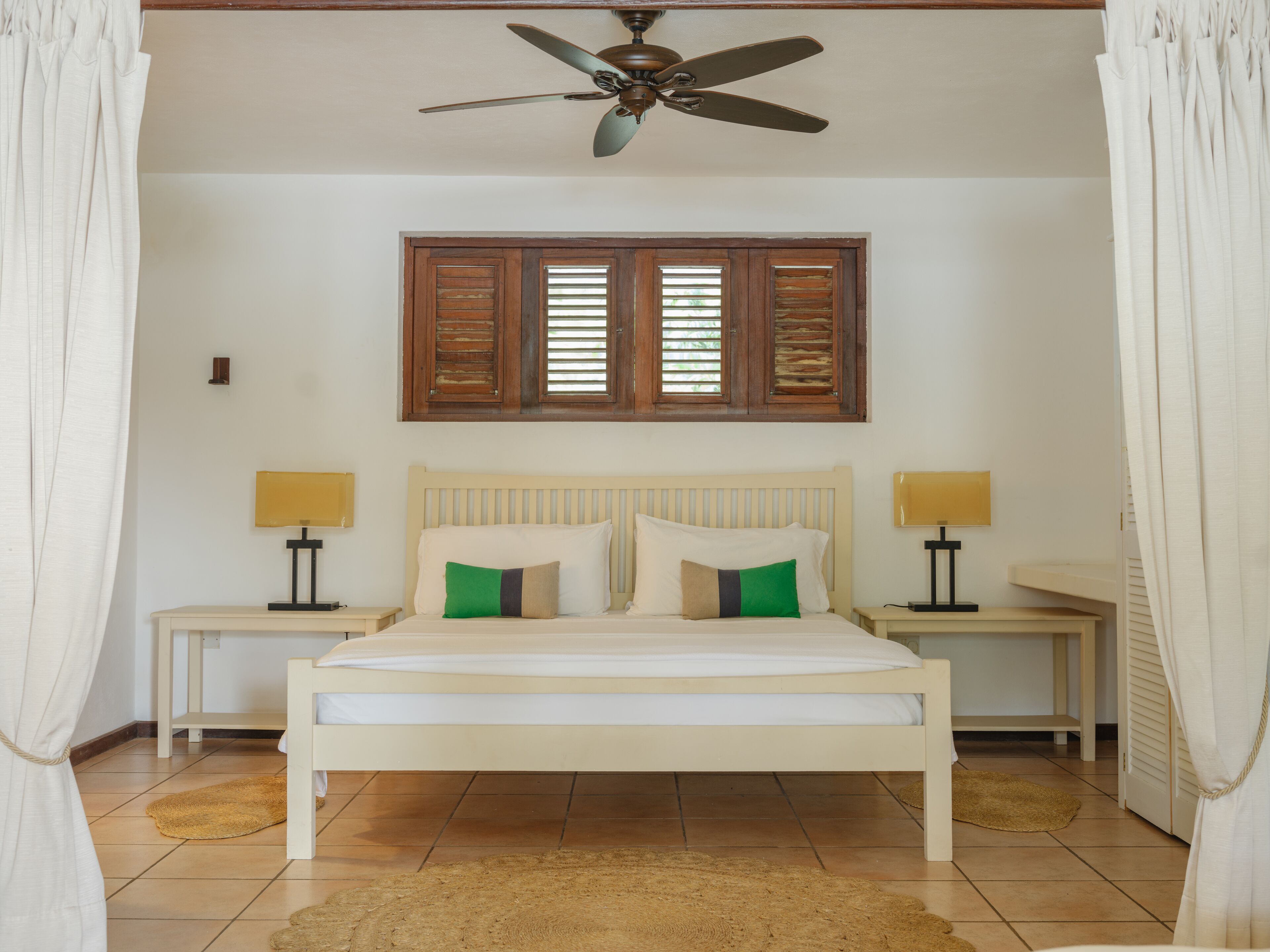 Une chambre à coucher avec un lit, deux tables de chevet, un ventilateur de plafond et une fenêtre avec des volets.