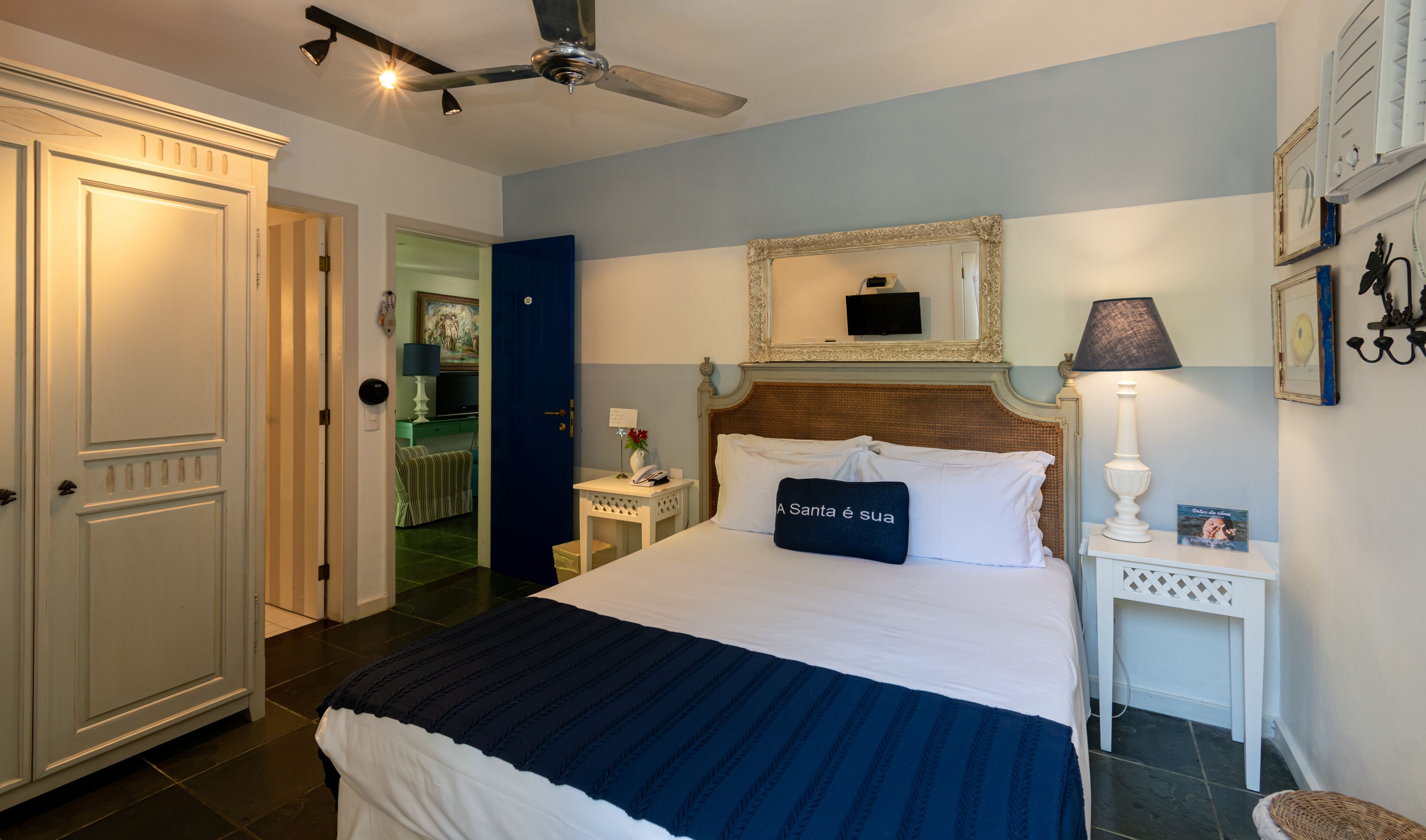 Habitación de hotel con cama, mesita de noche, espejo, ventilador de techo y puerta que da a otra habitación.