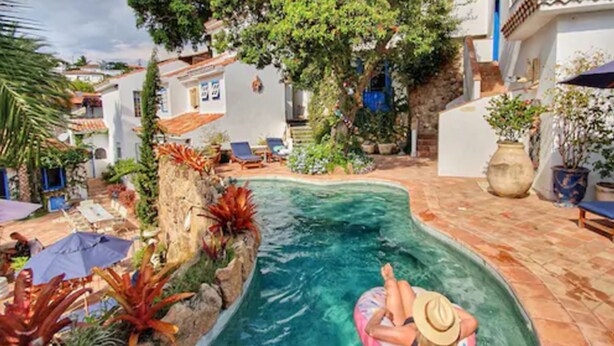 Una piscina con una persona en un flotador, rodeada de plantas y un muro de piedra.