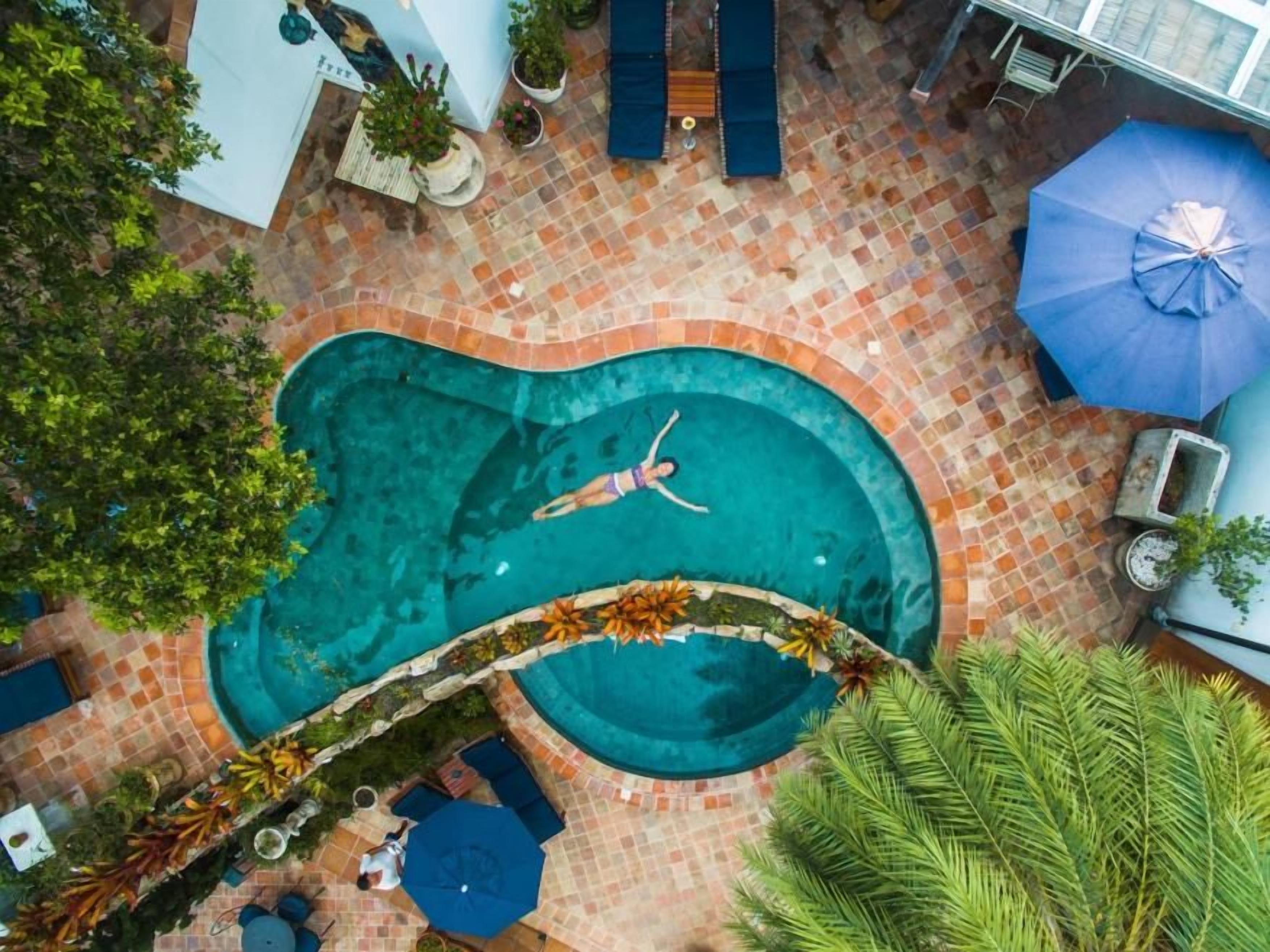 Ein Pool mit geschwungener Kante, umgeben von einer gefliesten Terrasse mit Liegestühlen und Sonnenschirmen.