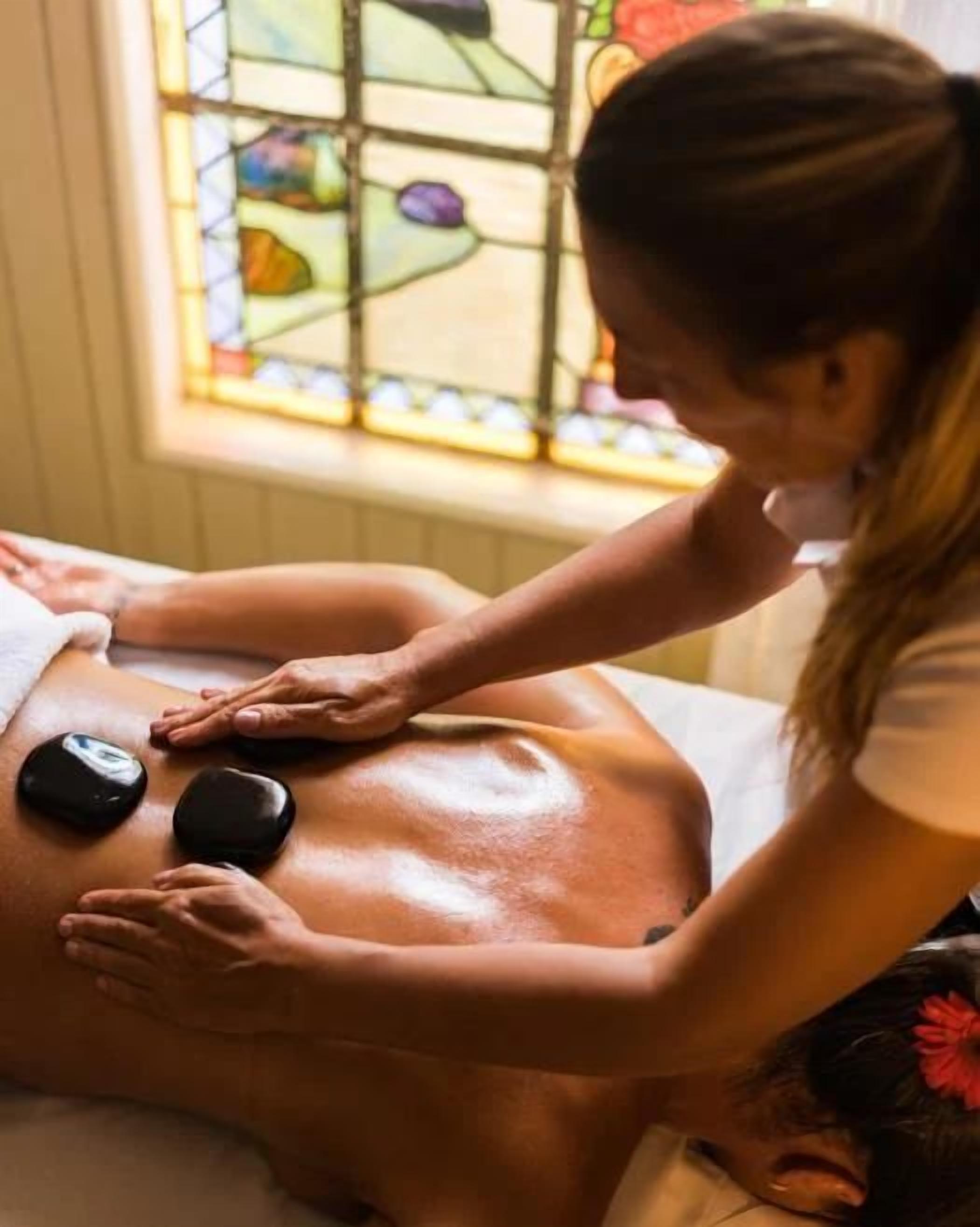 Sessão de massagem com uma pessoa deitada em uma mesa de massagem, um terapeuta aplicando pressão com as mãos e uma janela de vitral ao fundo.