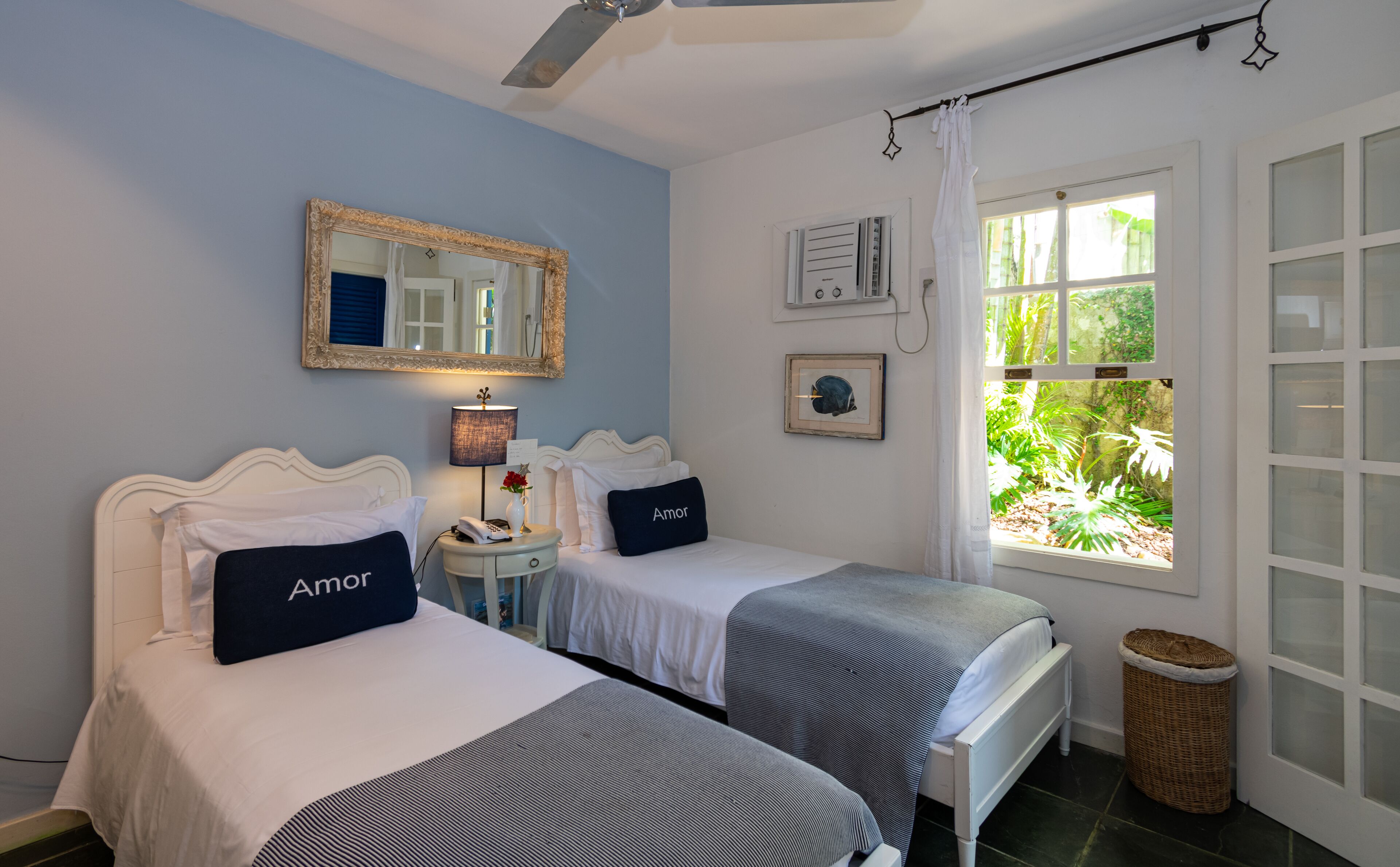 A bedroom with two beds, a mirror, a lamp, a window with a view of greenery, and a wicker basket.