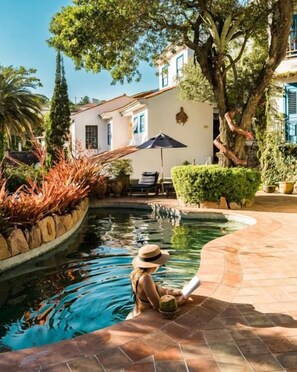 Un área de piscina con borde curvo, un patio con mesa y sombrilla, y un edificio con fachada blanca y detalles azules al fondo.
